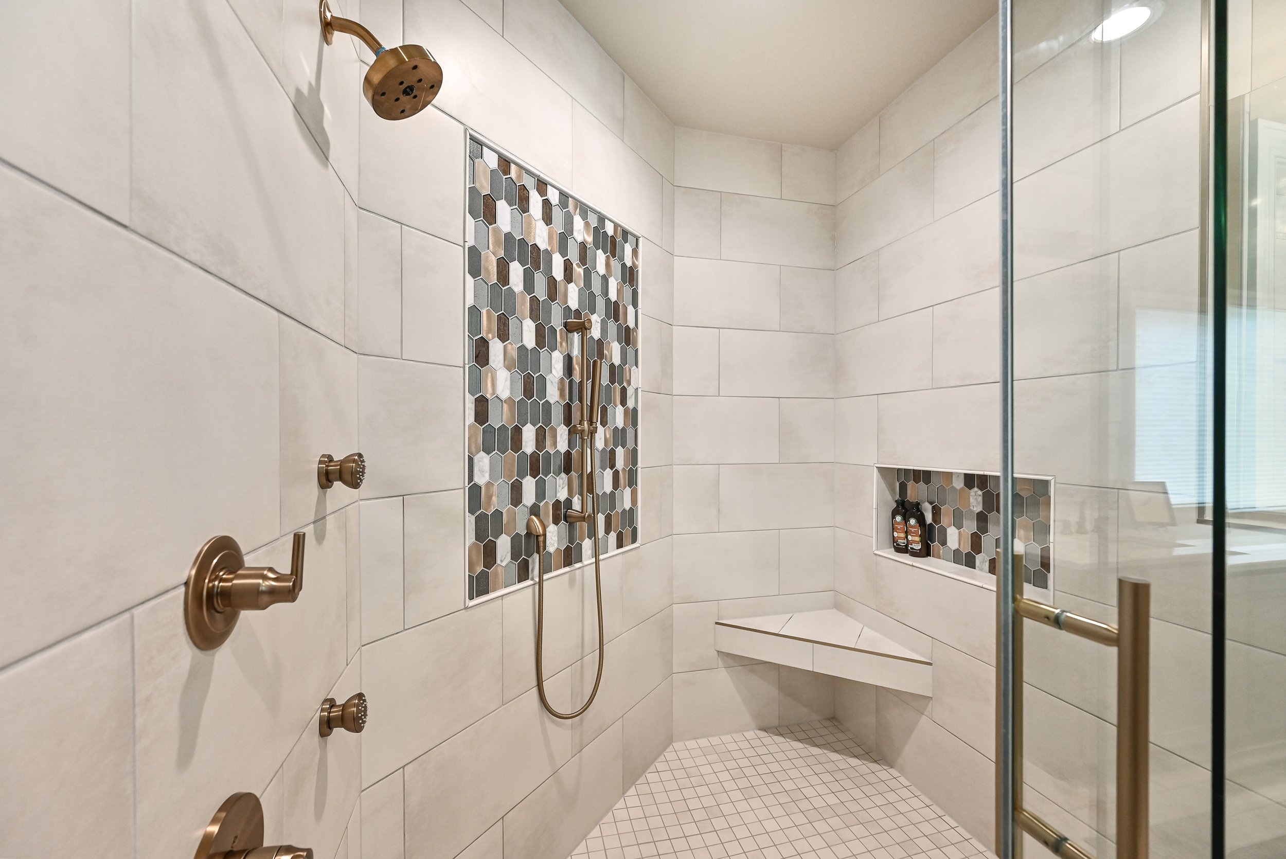 Modern walk-in shower with beige tile walls, a hexagon mosaic tile accent strip, and a built-in corner seat with toiletries, glass door, and bronze fixtures.