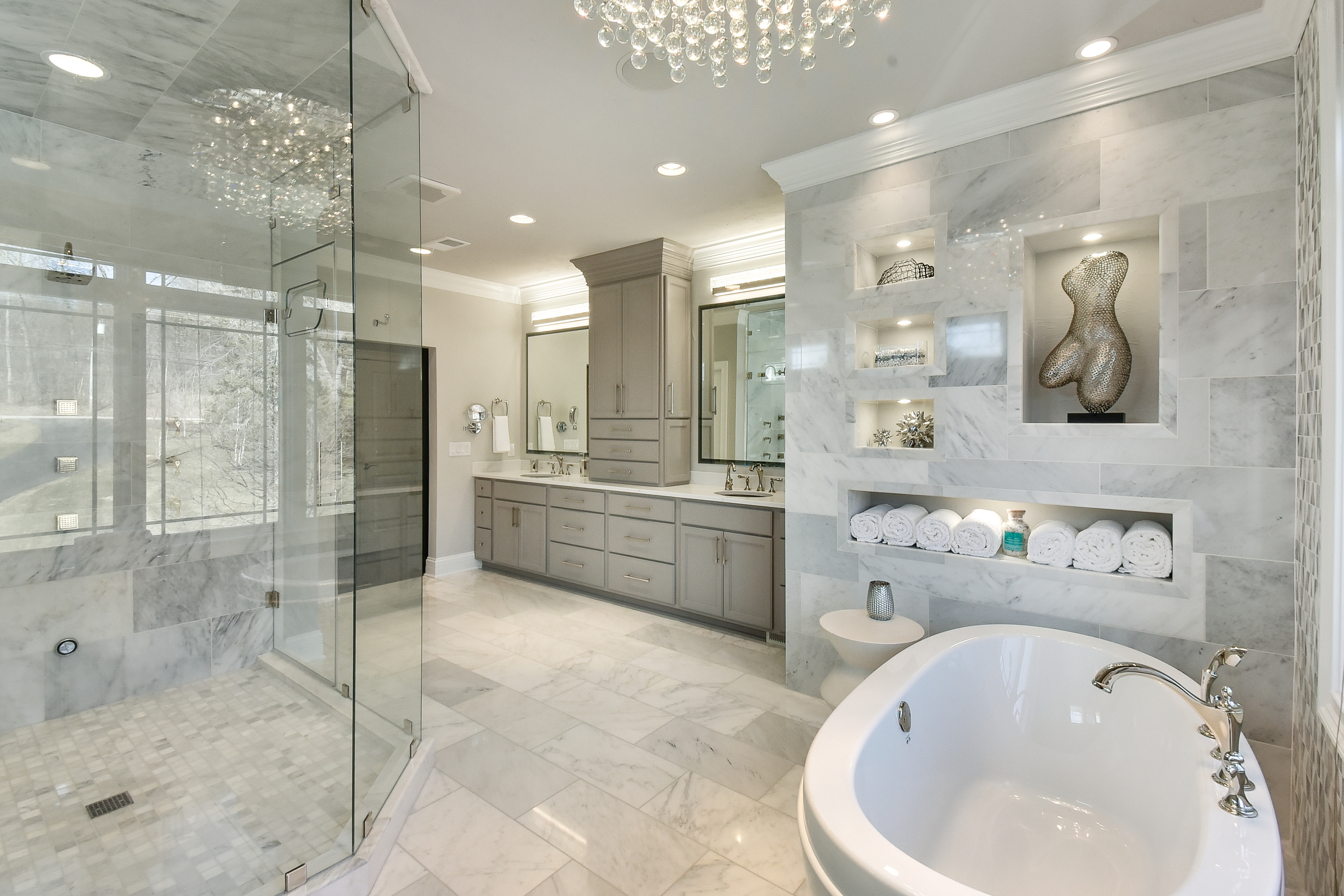Luxurious bathroom with a large glass-enclosed shower, a double vanity with two mirrors, a freestanding bathtub, marble walls and floors, built-in shelves with decorative items, and rolled white towels.
