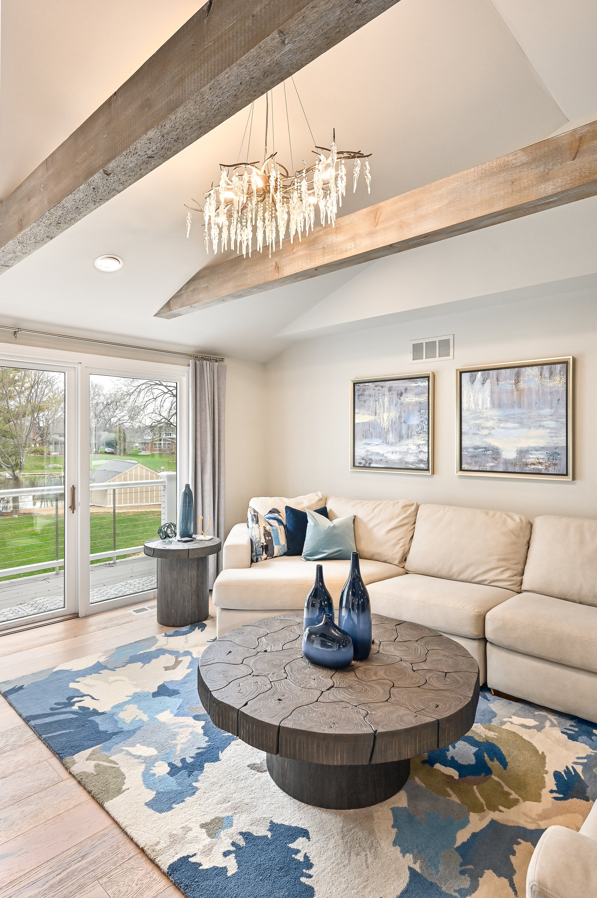 Living room with white sectional sofa, pressed wood coffee table with decorative vases, abstract paintings, sliding glass door leading outside, and chandelier hanging from wooden ceiling beams.