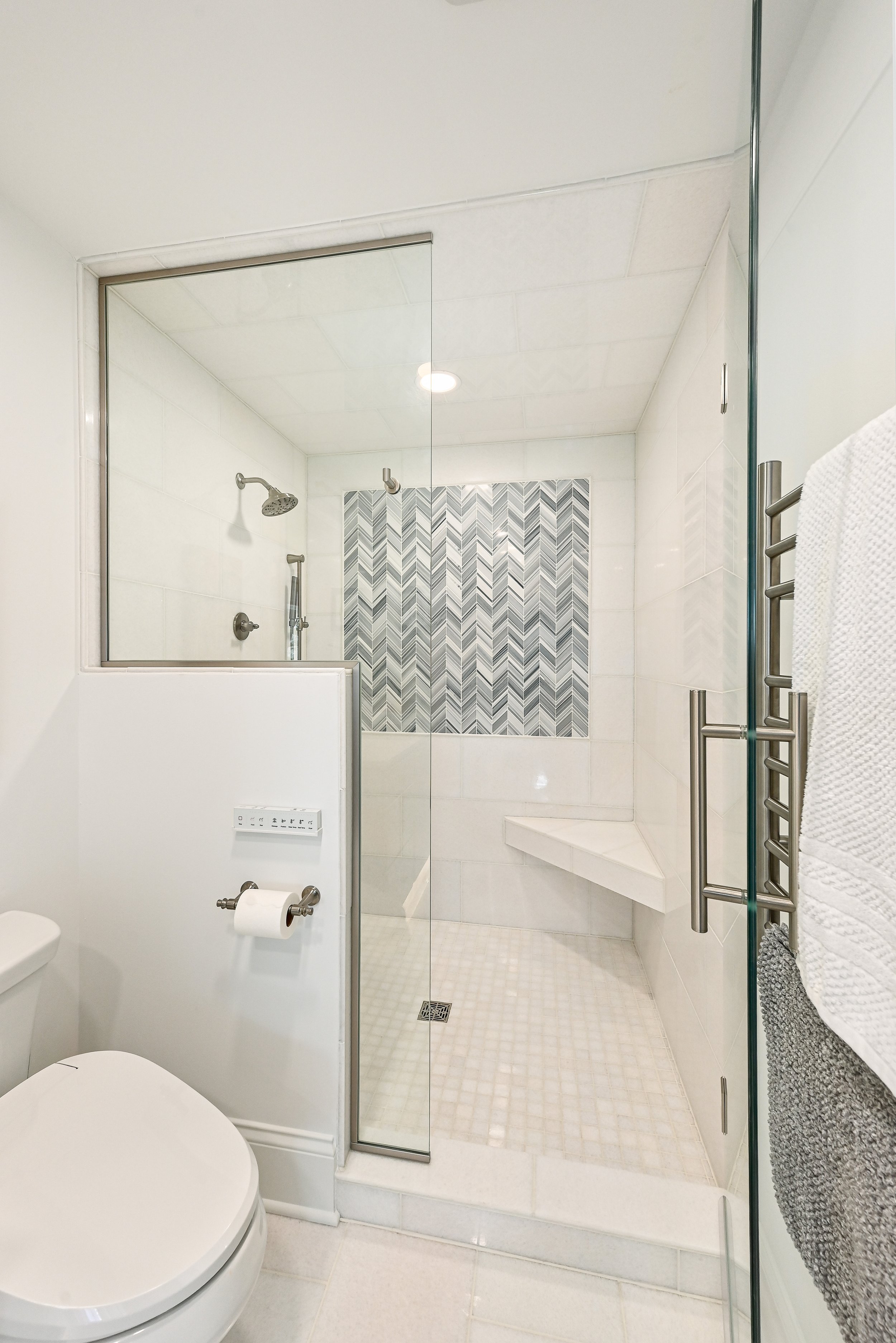 Modern bathroom with a walk-in shower featuring a gray chevron tile accent wall, glass enclosure, built-in bench, and white tiles.