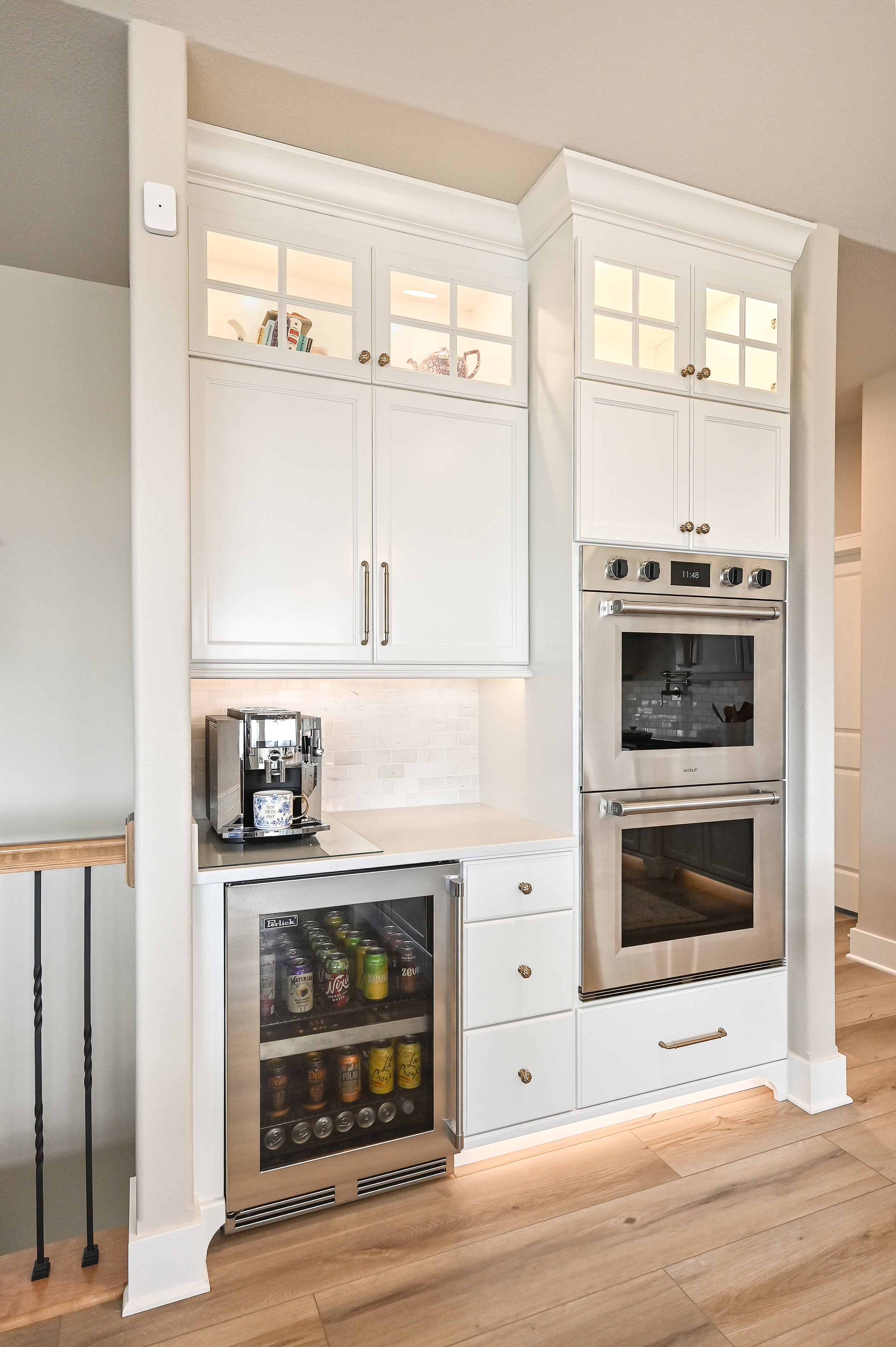 Kitchen with white cabinets, built-in double oven, mini fridge with drinks, coffee machine, and decorative cabinets.