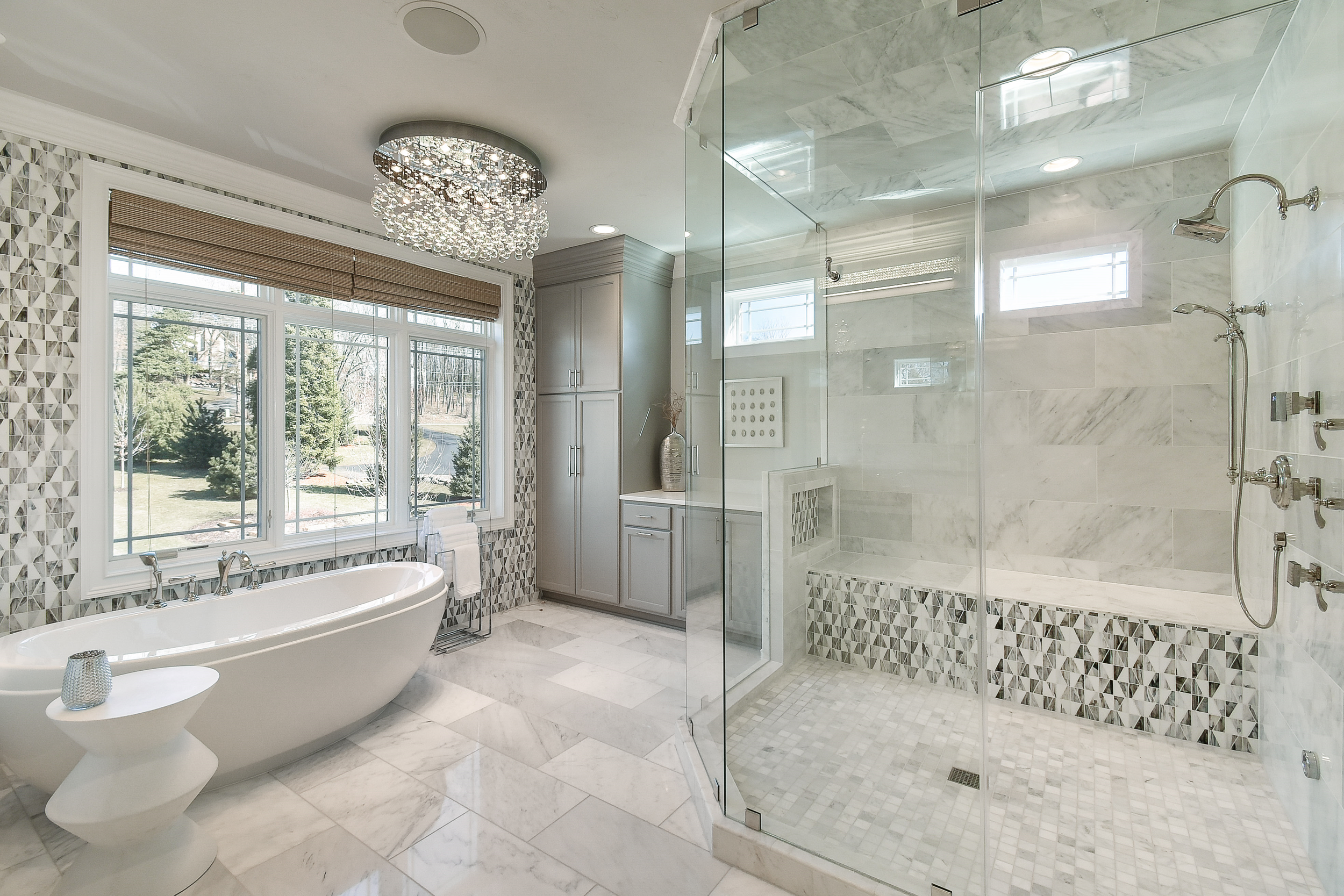 Modern bathroom with marble flooring, large window, freestanding bathtub, and a glass-enclosed shower with marble tile and multiple shower heads.
