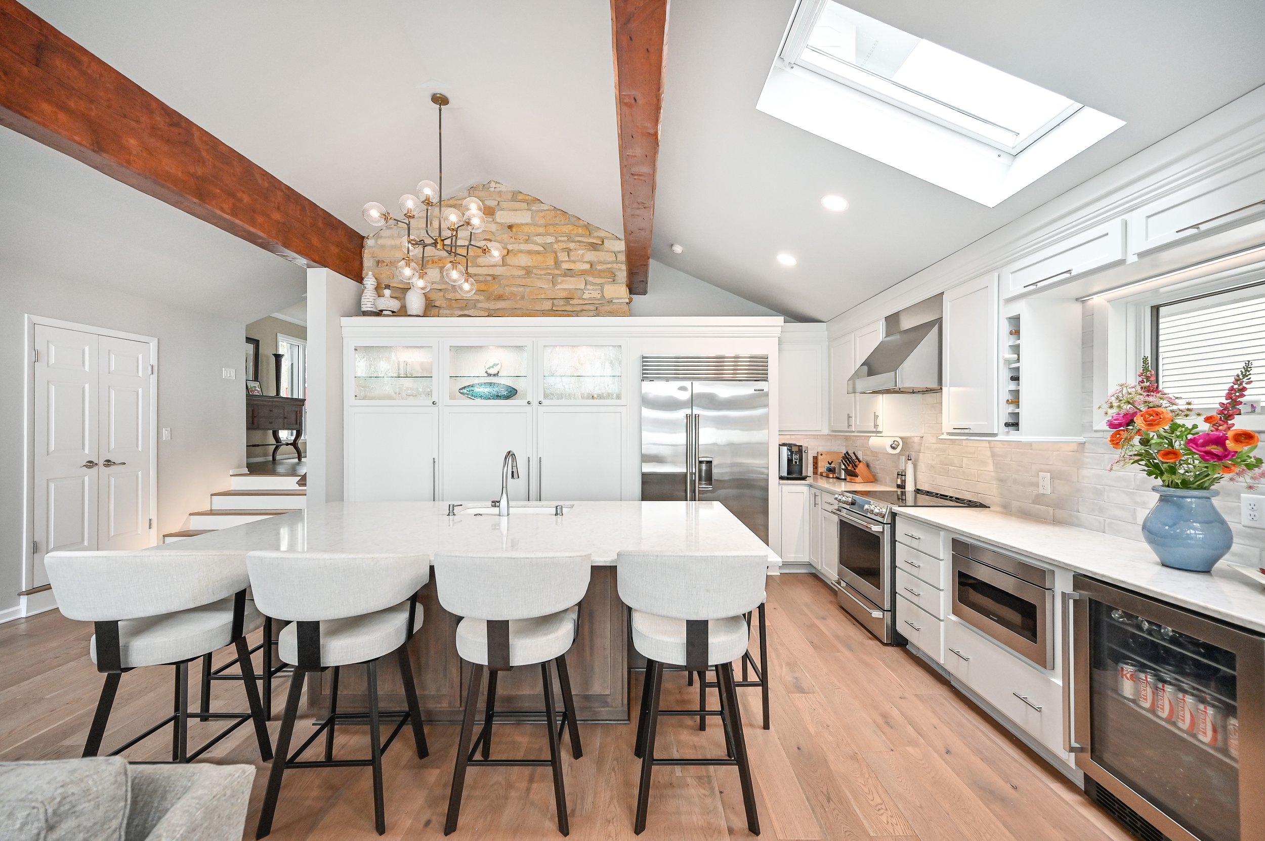 Modern kitchen with white cabinets, stainless steel appliances, a large island with bar stools, and a skylight overhead. Decor includes a flower vase on the countertop.