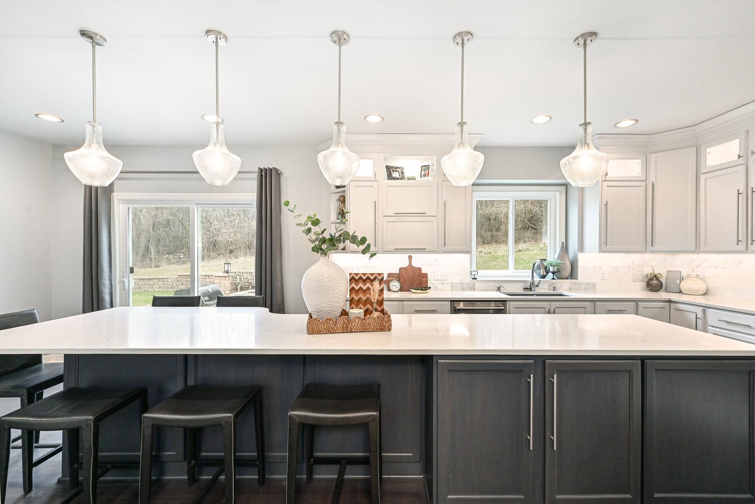 Modern kitchen with white countertops, gray cabinets, black bar stools, pendant lights, and large windows showing an outdoor view.