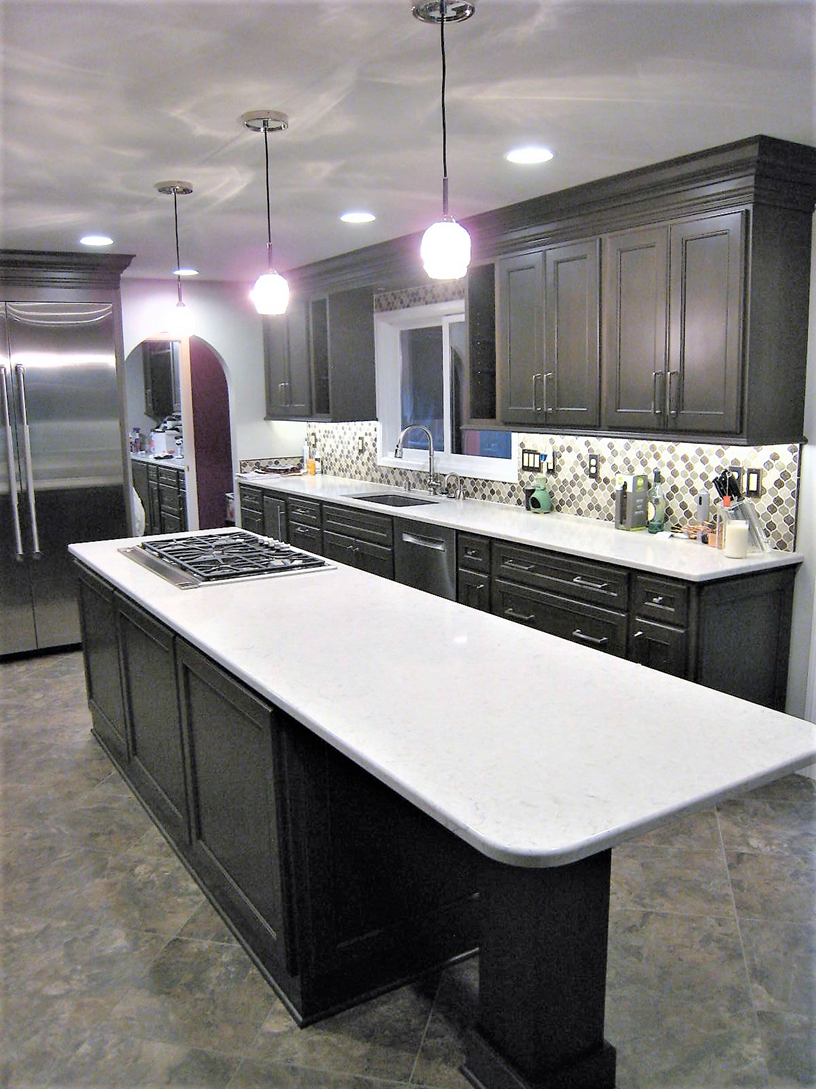 A modern kitchen with black cabinets, white countertops, a large kitchen island with a stove, and a backsplash with multicolored tile patterns. There are pendant lights hanging from the ceiling, a window over the sink, and stainless steel appliances.