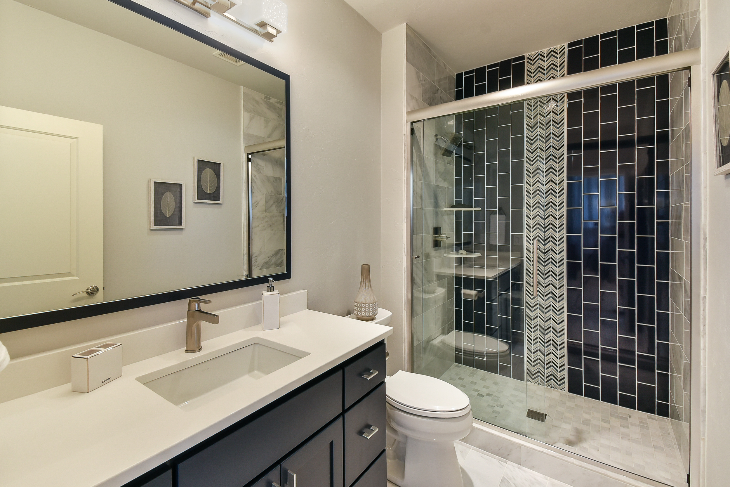 Modern bathroom with a white vanity, large mirror, and black cabinets. Shower with glass door and dark geometric tiles on the wall.