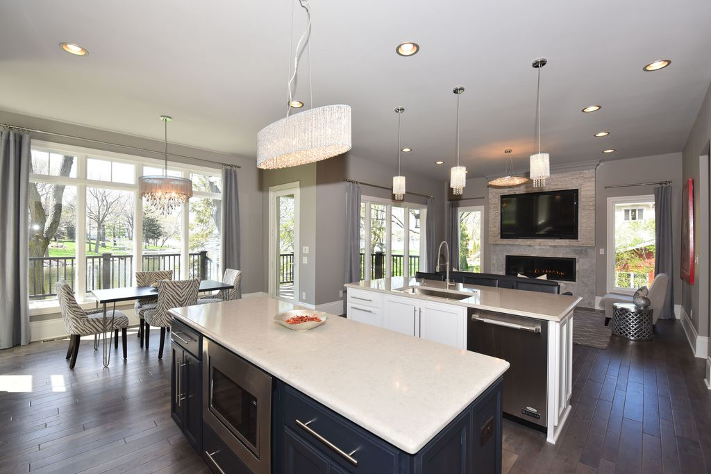 Open-concept kitchen and living room with large windows, gray walls, a white island, a breakfast area with patterned chairs, a modern fireplace, and a flat-screen TV.