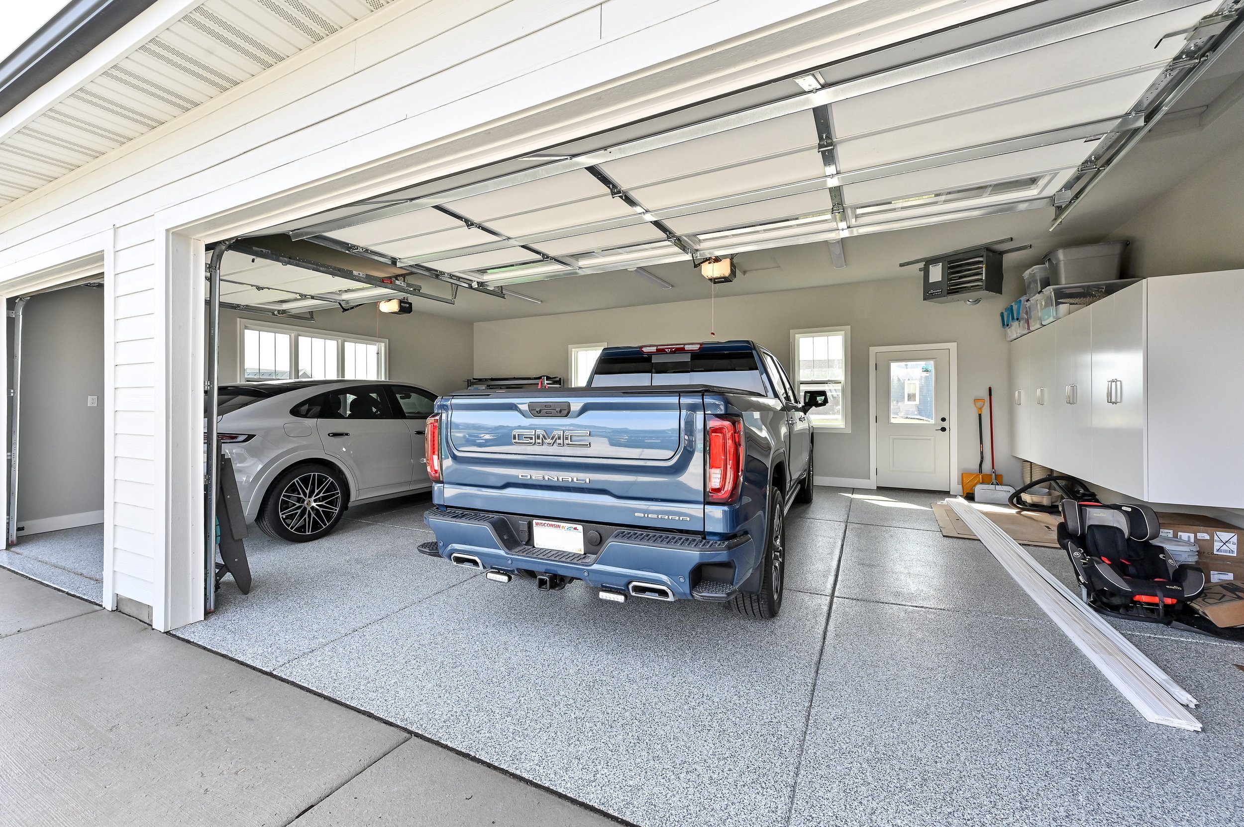 A garage with a blue GMC Sierra truck in the center, and a gray car parked on the left side. The garage has a speckled gray floor, white walls, and an open wide door. There are various storage cabinets, tools, and a child's car seat on the right side
