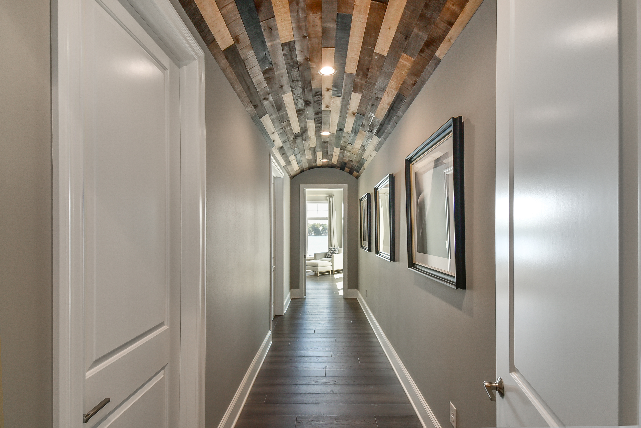 A narrow hallway with gray walls, dark hardwood flooring, and a wooden arched ceiling with recessed lighting. Framed artwork decorates the right wall, and at the far end, there's a seating area with a white chair and a window showing outdoor scenery.