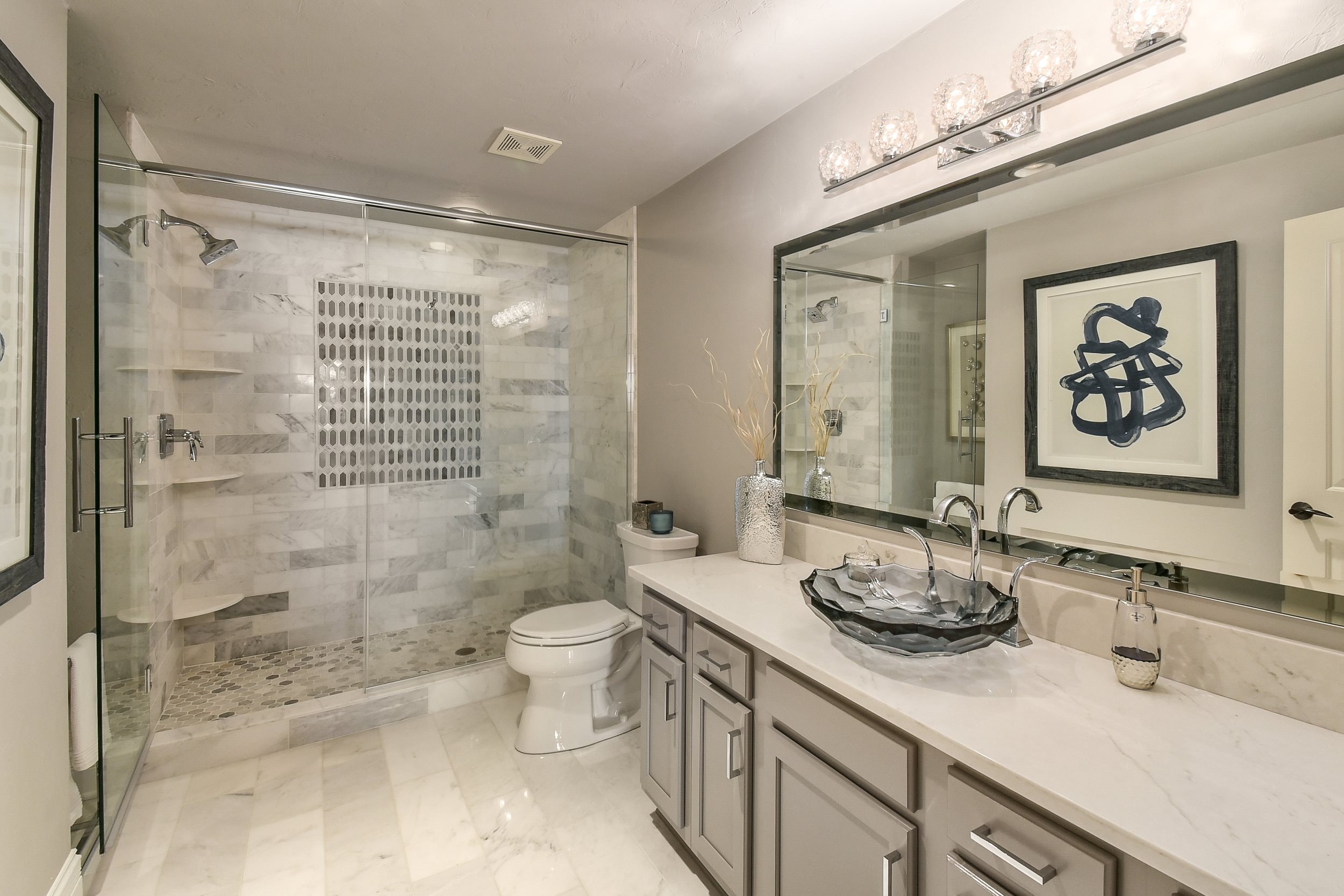 A modern bathroom featuring a glass-enclosed shower with marble tiles, a white toilet, a beige vanity with a decorative sink, and a large mirror highlighted by light fixtures. Decor includes a framed abstract artwork, vases, and a soap dispenser.
