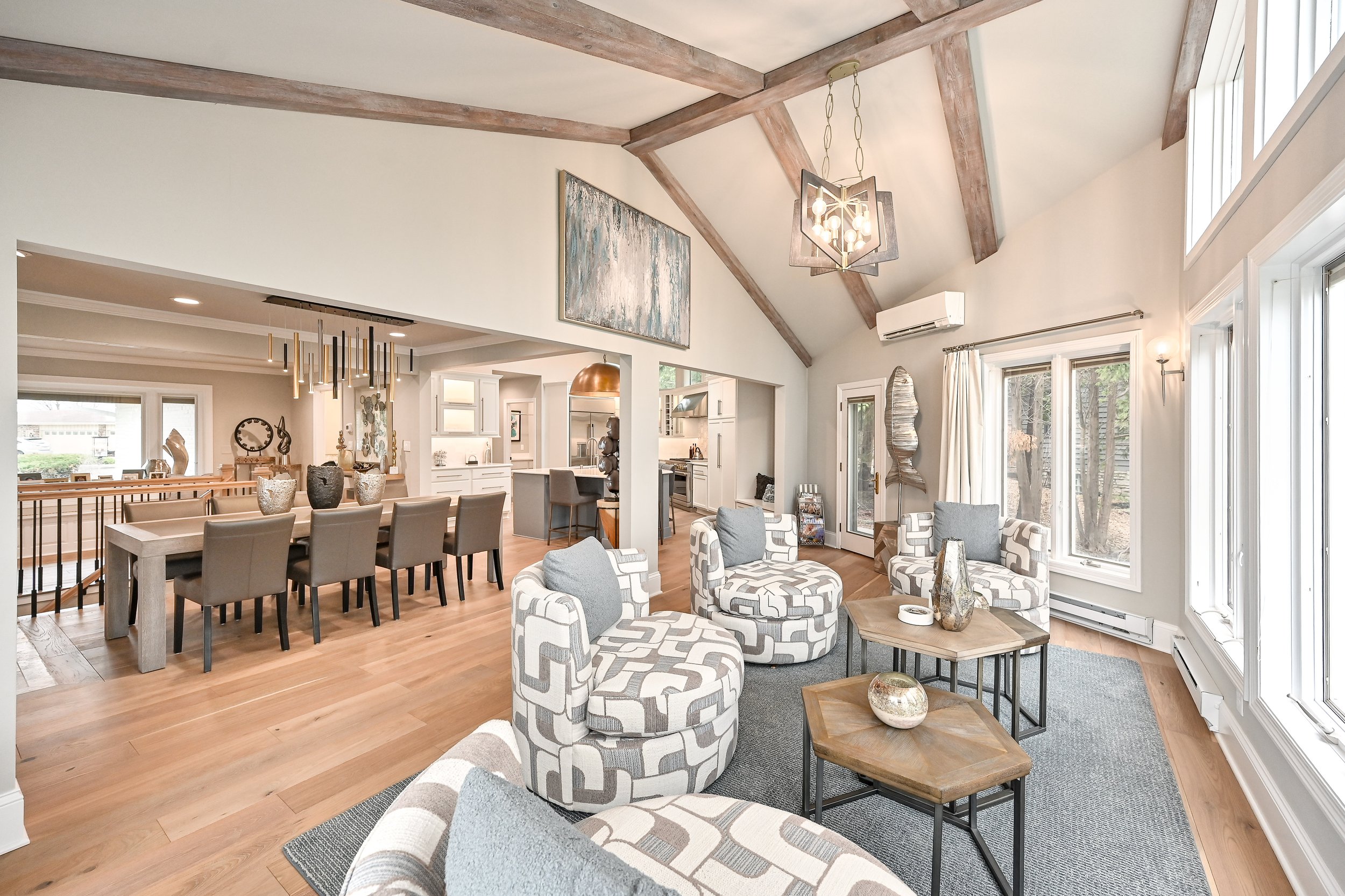 Open-concept living and dining room with vaulted ceiling, wooden beams, large windows, and modern decor, including patterned armchairs, a chandelier, and decorative vases.