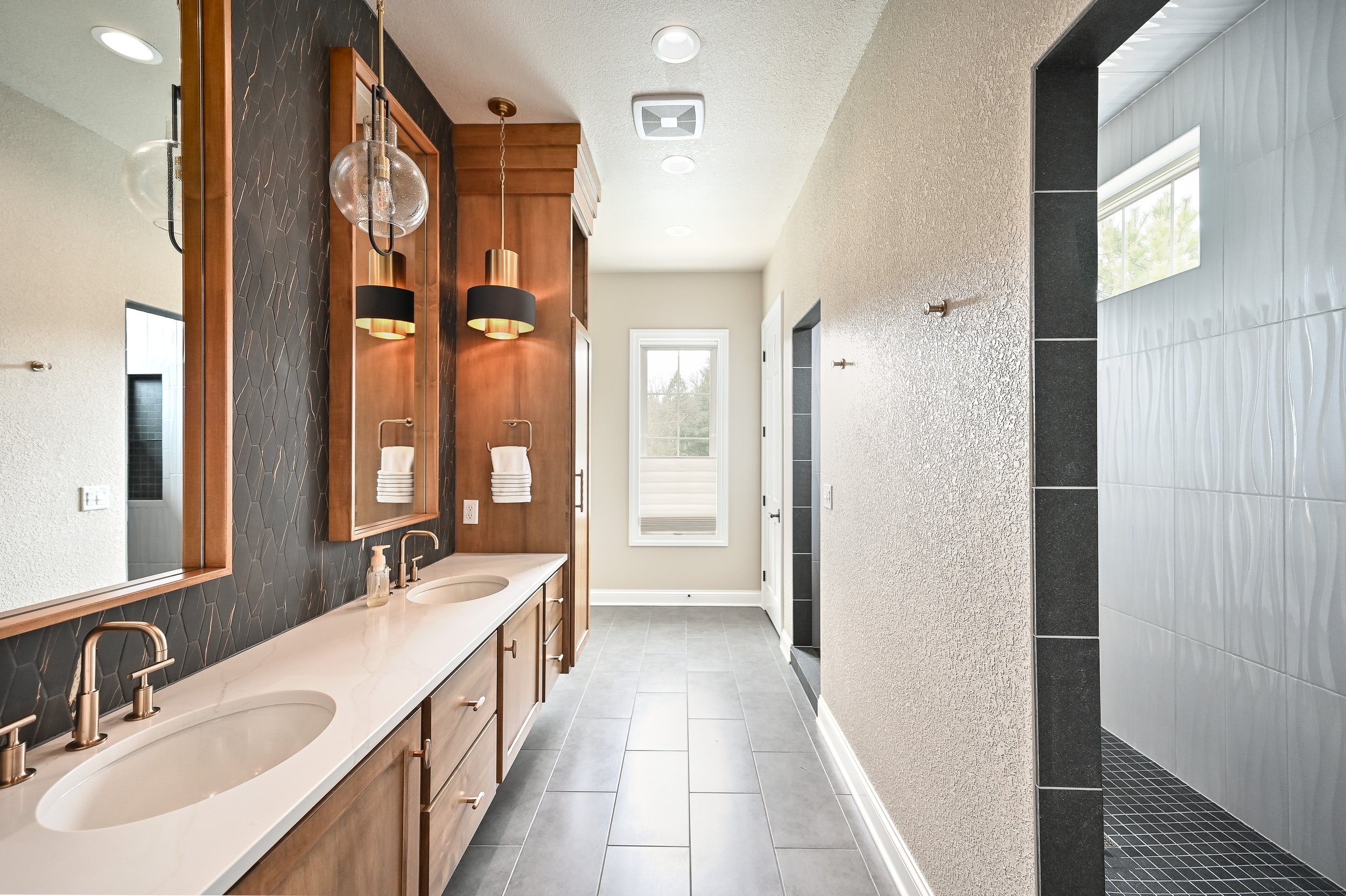 Modern bathroom with dual vanity sinks, black textured accent wall, wood cabinetry, hanging pendant light, wall-mounted towel ring, and a walk-in shower with gray and black tile.