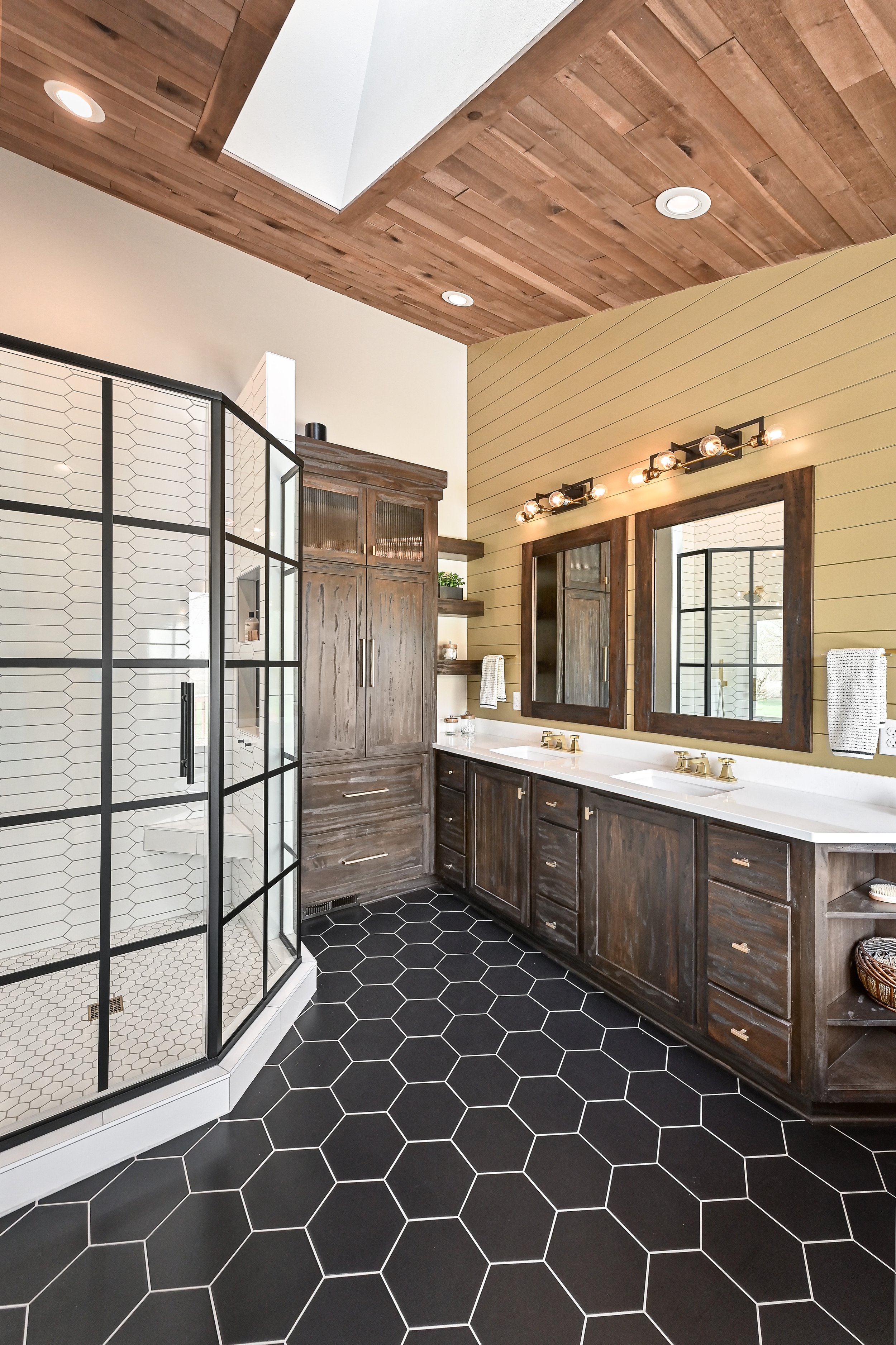 A modern bathroom featuring a black hexagonal tile floor, a large mirror with a wooden frame above a double sink vanity, wooden cabinetry, open shelves, a walk-in shower with black-framed glass doors, a wood-paneled ceiling, and beige painted walls.