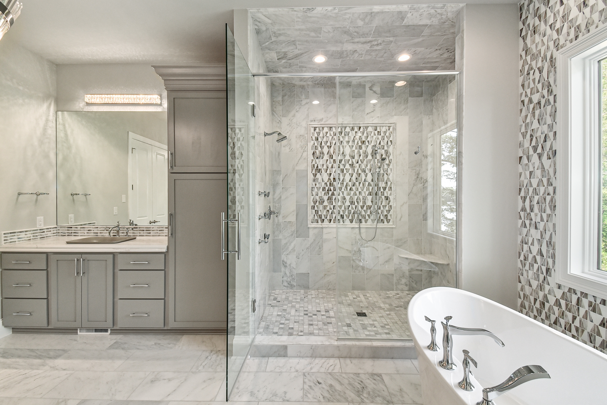 A modern bathroom with a glass-enclosed shower, a large bathtub with chrome fixtures, a grey vanity with a mirrored wall, and patterned tiles around the shower and on the wall near the window.