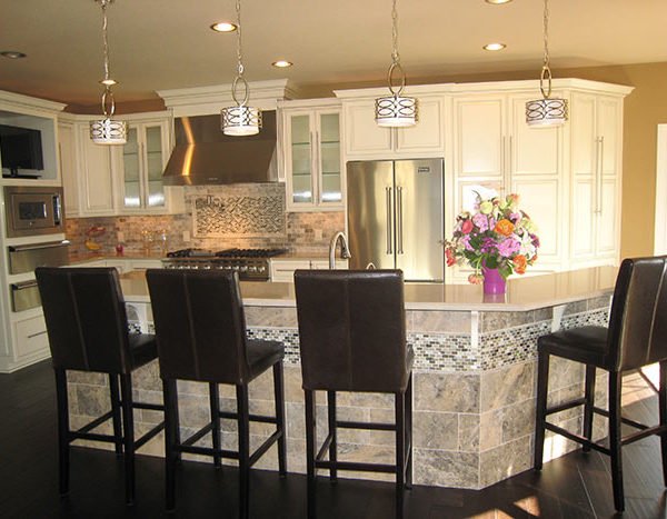 Modern kitchen with white cabinets, stainless steel refrigerator, and a curved island with four black barstools. Pendant lights hang above the island, and a colorful flower arrangement is on the counter.