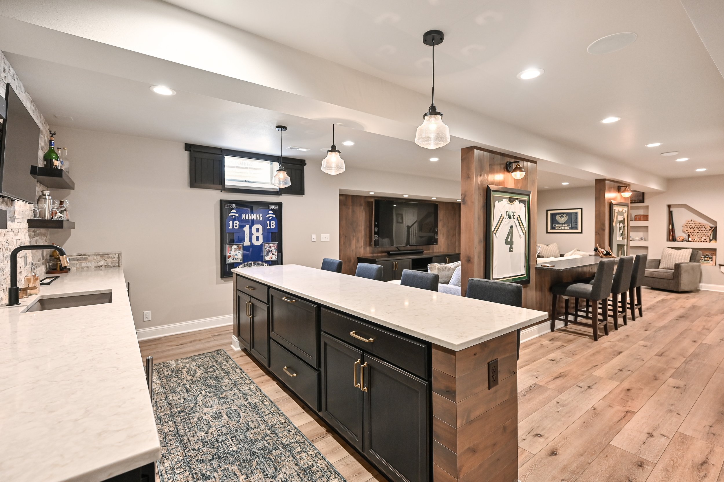 A spacious open-concept basement with a kitchen, bar, and seating area. The kitchen has white countertops, black cabinetry, and pendant lighting. The bar area has high chairs, and the living space features framed sports jerseys and a TV.