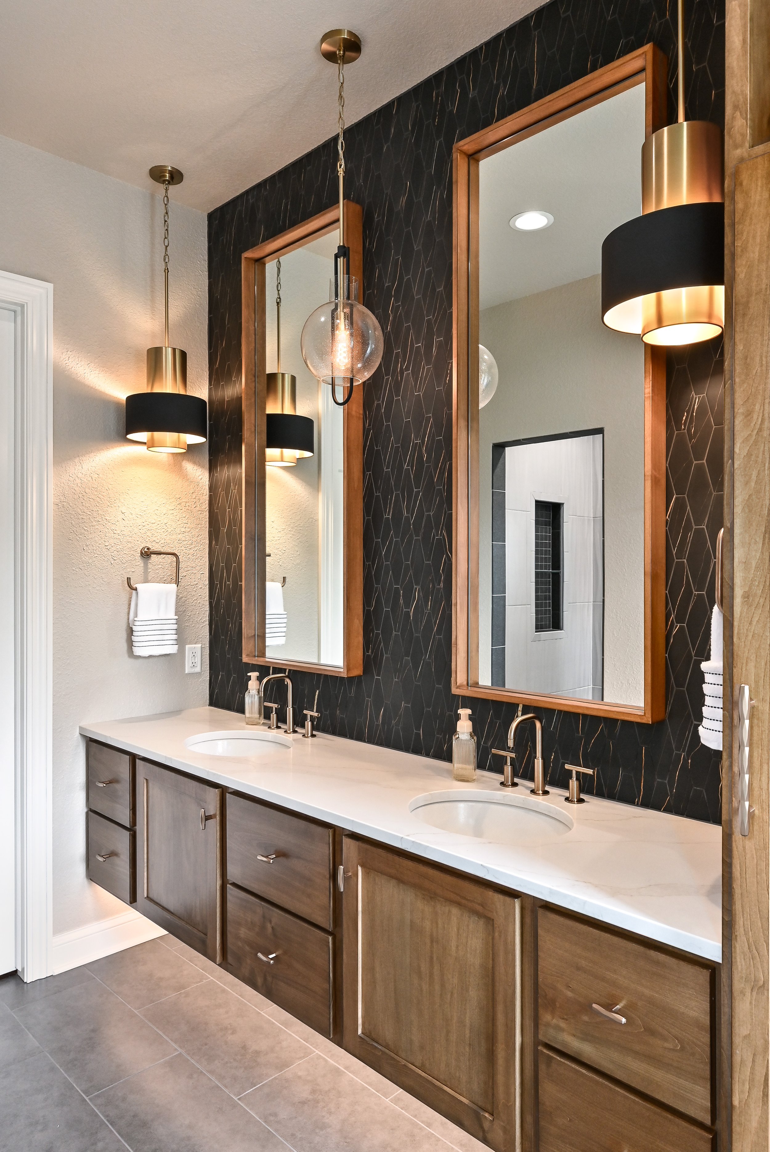 Bathroom vanity with two sinks, wooden cabinets, white countertop, two mirrors, black and gold wall sconces, modern pendant light, and neutral wall and tile decor.