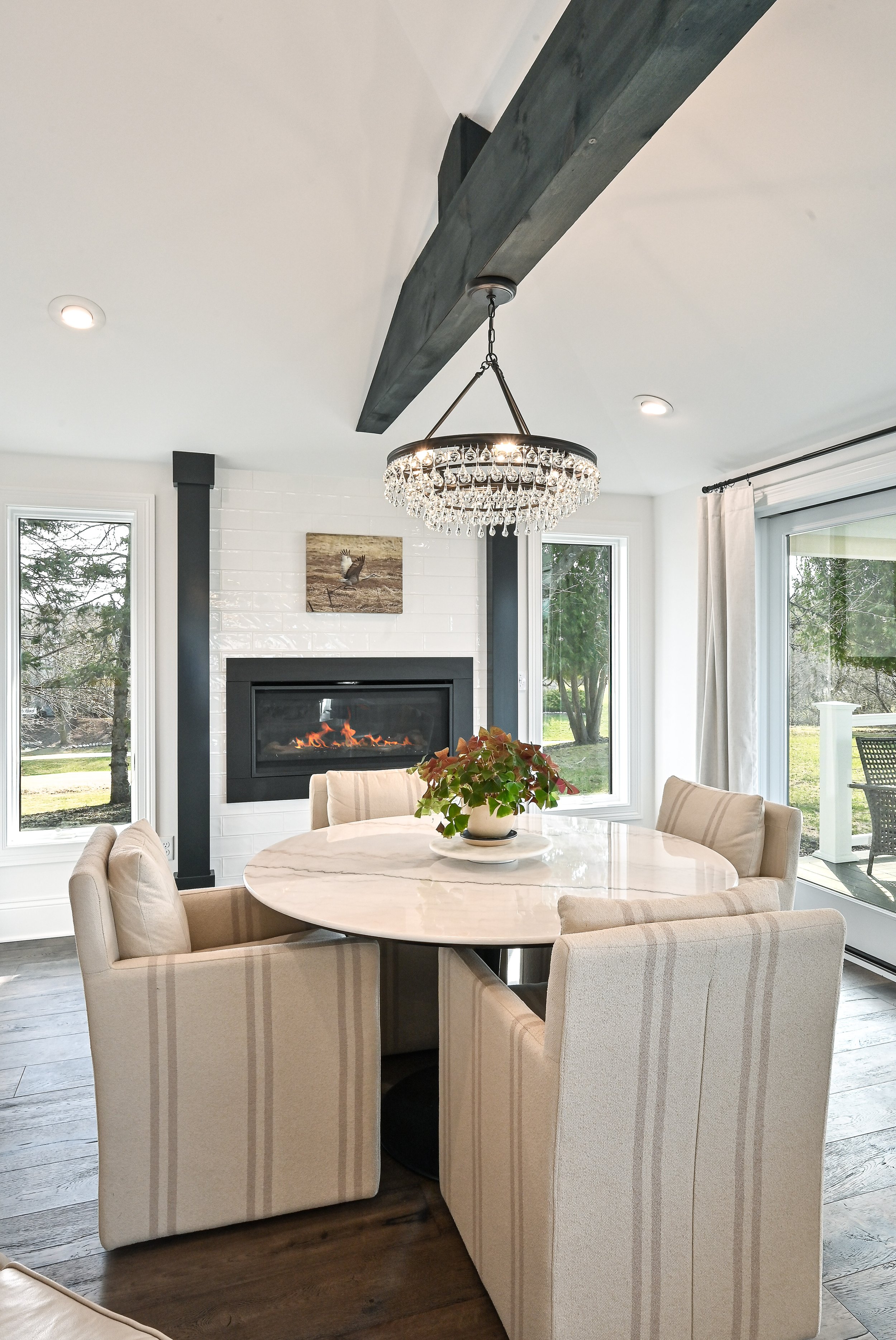 A dining room with a round white table, beige upholstered chairs, a chandelier, a fireplace, and large windows showing an outdoor patio and trees.