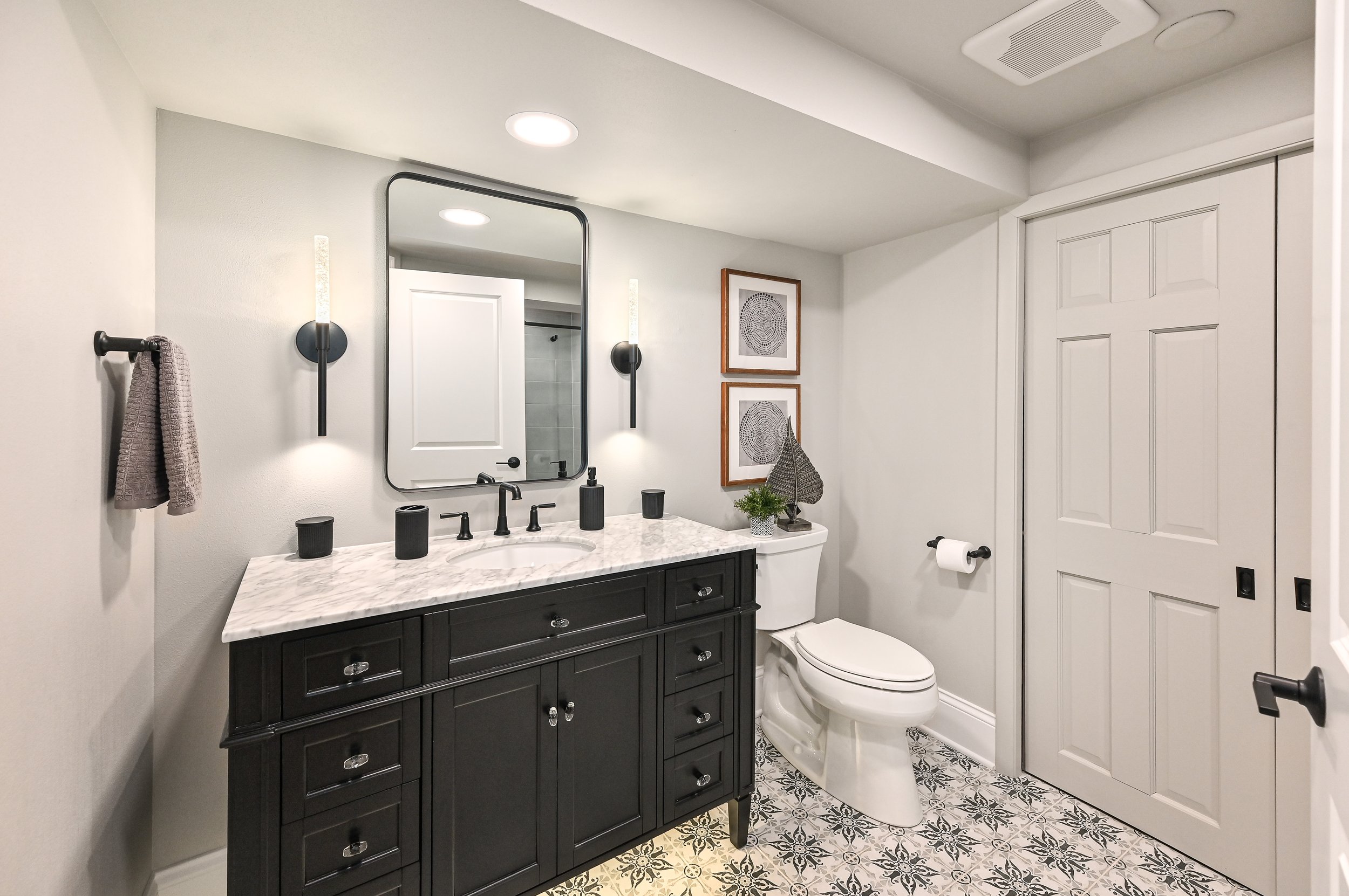 Modern bathroom with black vanity, marble countertop, mirror, wall sconce, framed artwork, plant, and toilet, with patterned tile floor and closed closet doors.