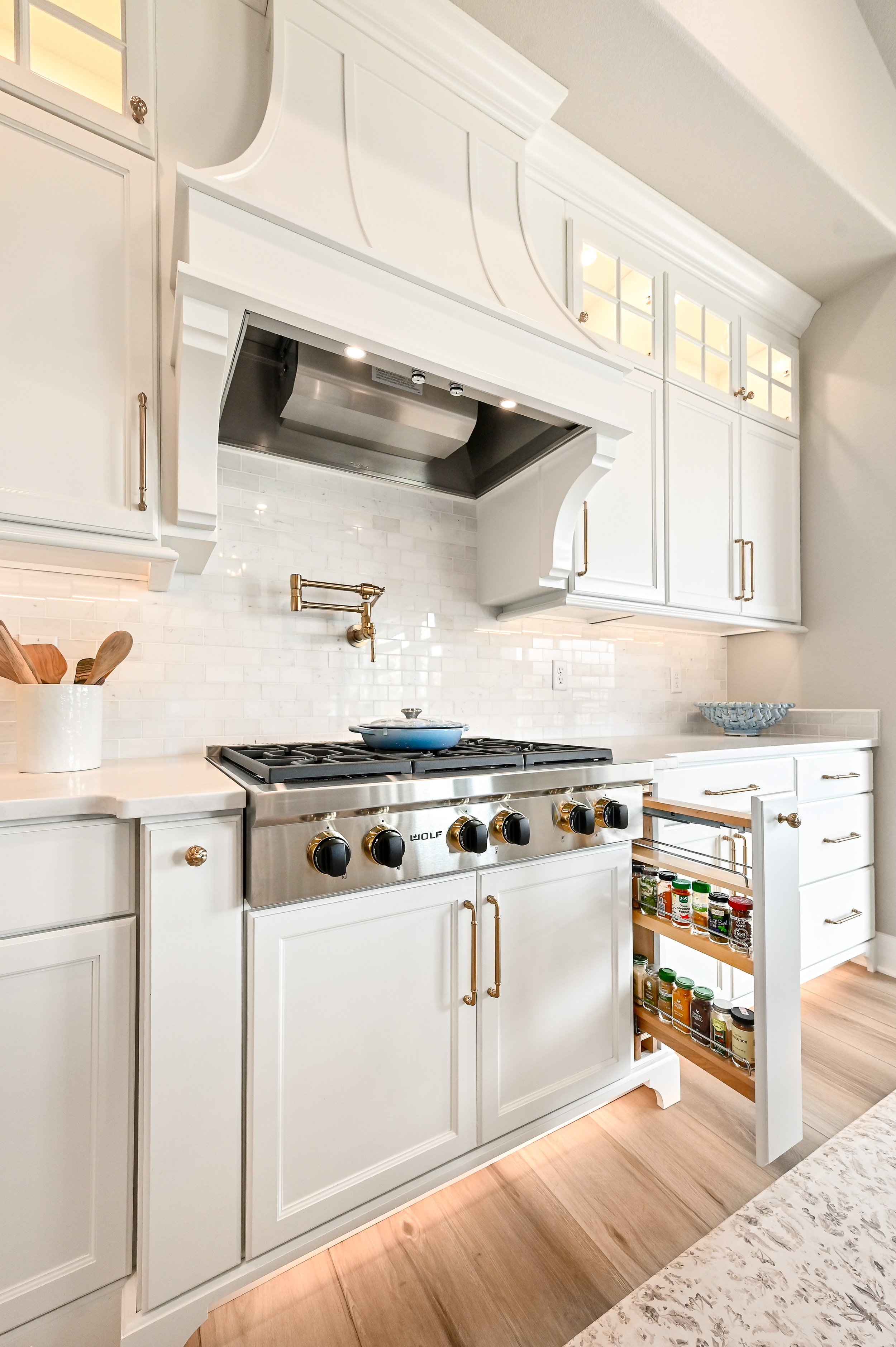 A white kitchen with wood flooring, white cabinetry, a stainless steel stove with black and gold knobs, and a brass pot filler faucet above the stove. There is a spice rack drawer on the right side and a decorative bowl on the countertop.