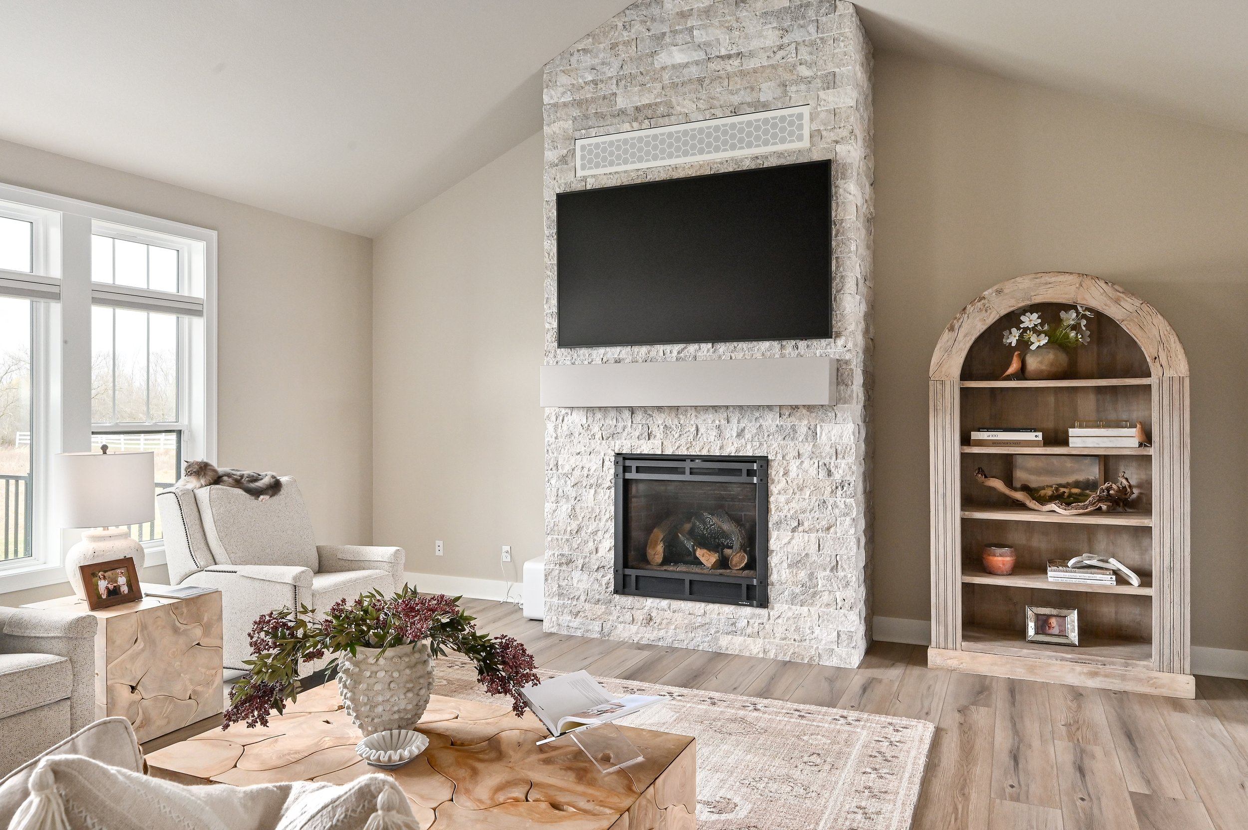 Living room with a large window, beige couches, a wooden coffee table with a vase of flowers, a stone fireplace with a TV mounted above, and a wooden bookshelf with decor and books.