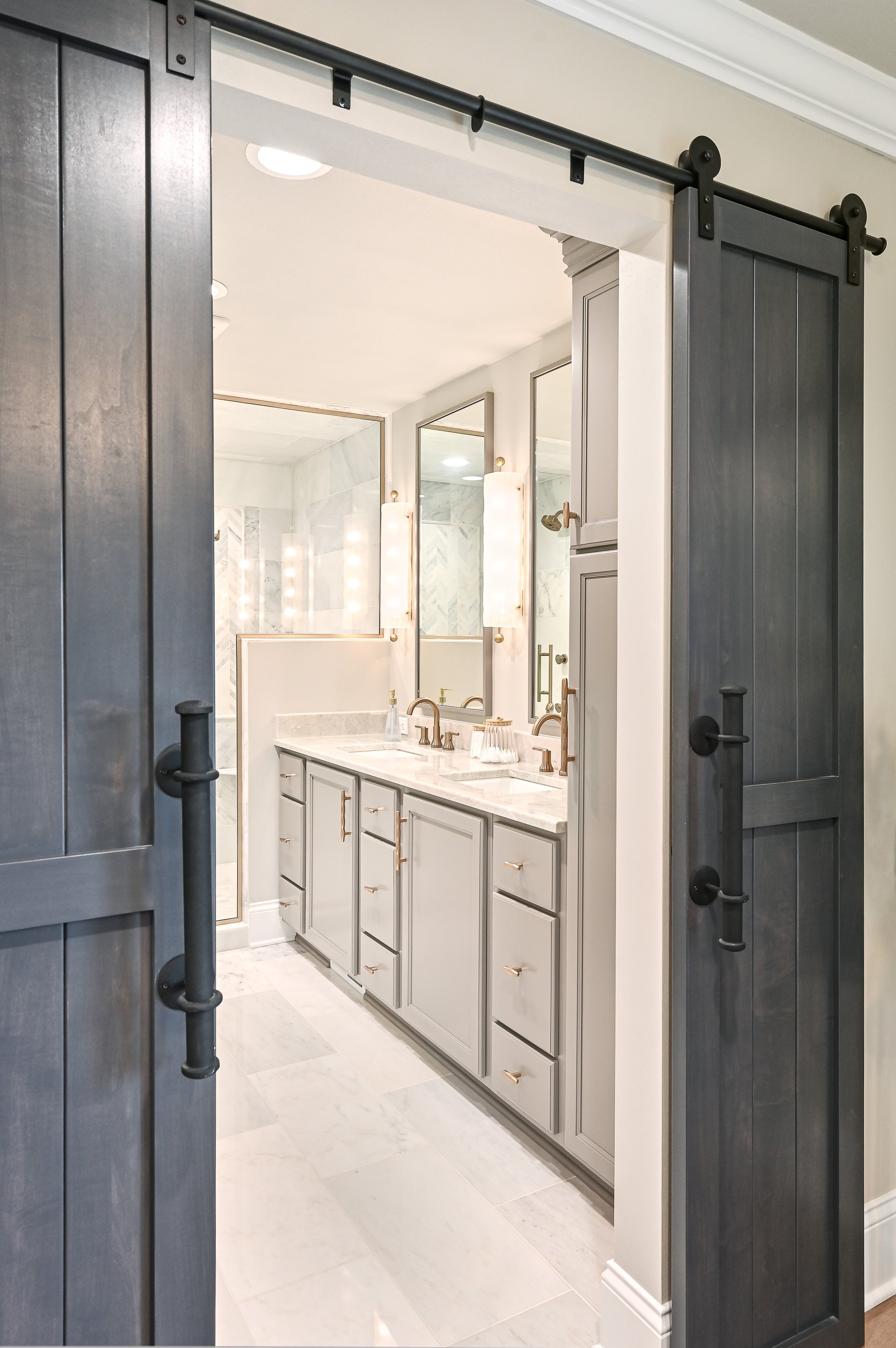 View of a modern bathroom through sliding barn doors, featuring a double vanity with light gray cabinets, a white marble countertop, and two large mirrors with wall-mounted light fixtures.