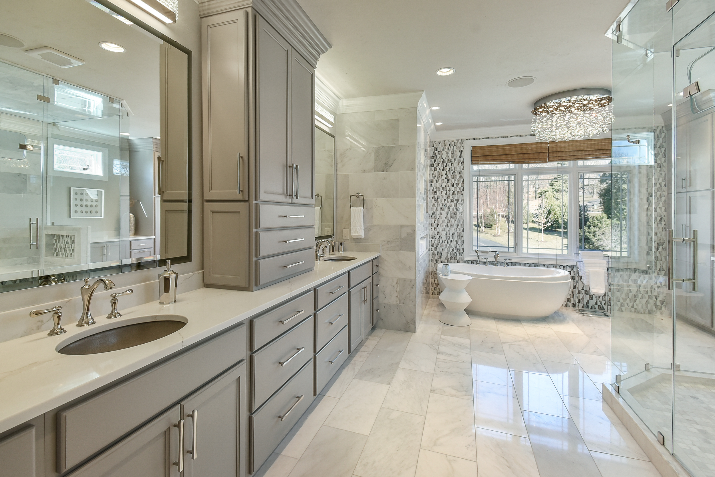Modern bathroom with gray cabinetry, marble flooring, a bathtub by large windows, a glass shower, and a crystal chandelier.