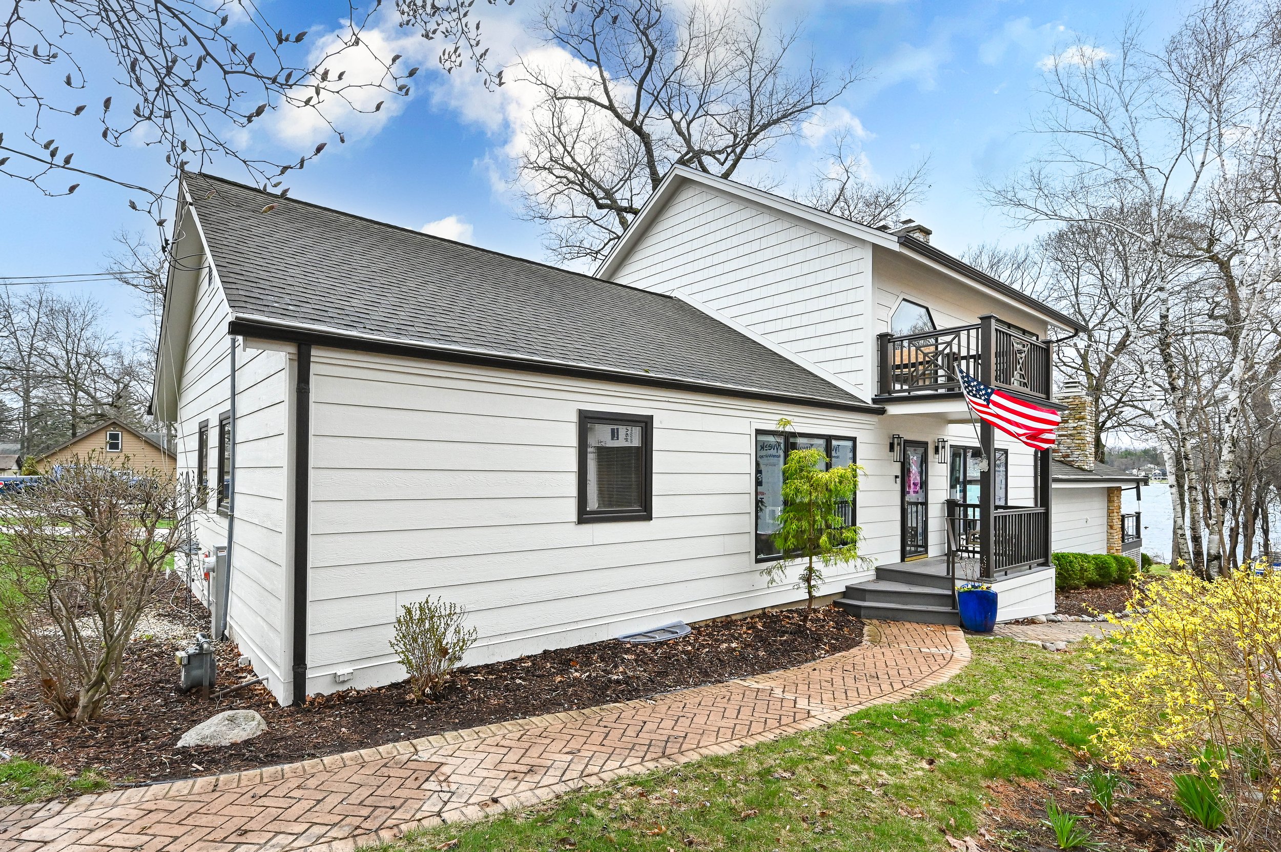View of a two-story white house with a black roof, black window frames, and black railings on a small balcony. An American flag is hanging from the balcony. The house has a brick walkway leading to the front door, surrounded by small trees and shrubs
