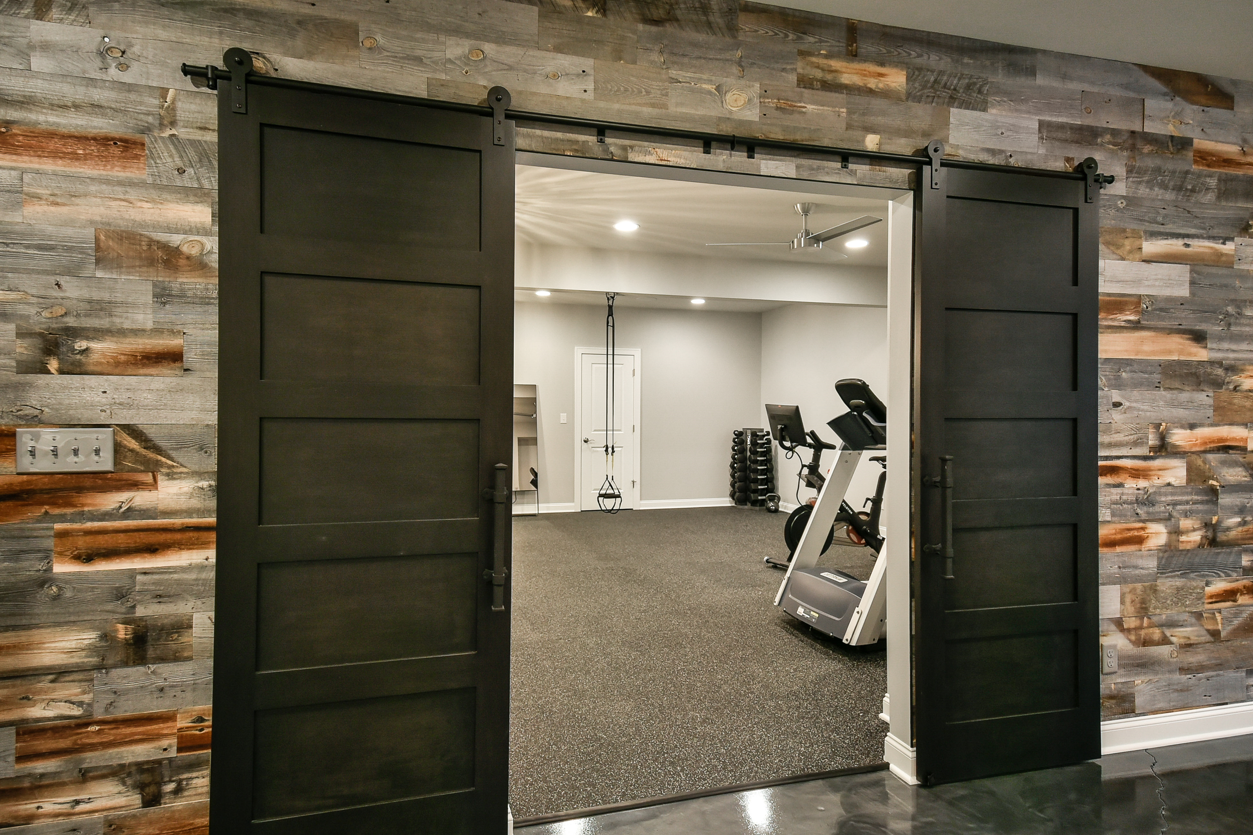 View of a home gym with black barn doors open, revealing exercise equipment like a stationary bike, dumbbells, and a suspension trainer inside a room with gray walls and dark carpet.