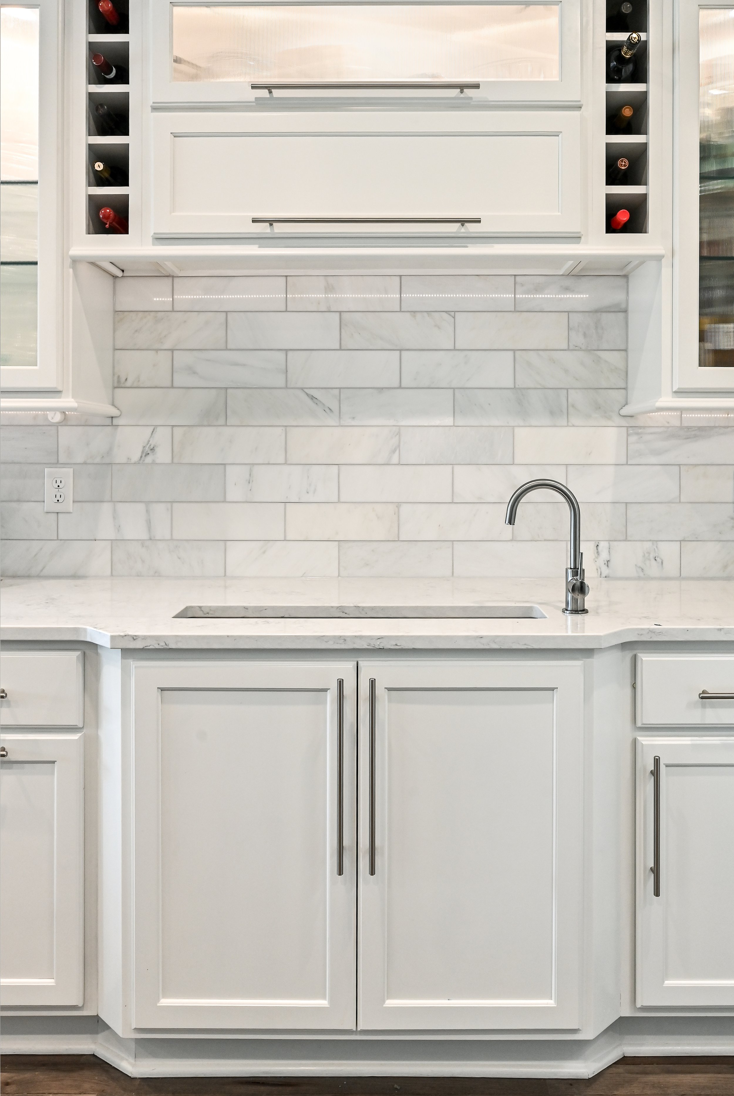 White kitchen with marble backsplash, undermount sink, stainless steel faucet, and white cabinetry, with wine bottles stored on either side of an upper cabinet.