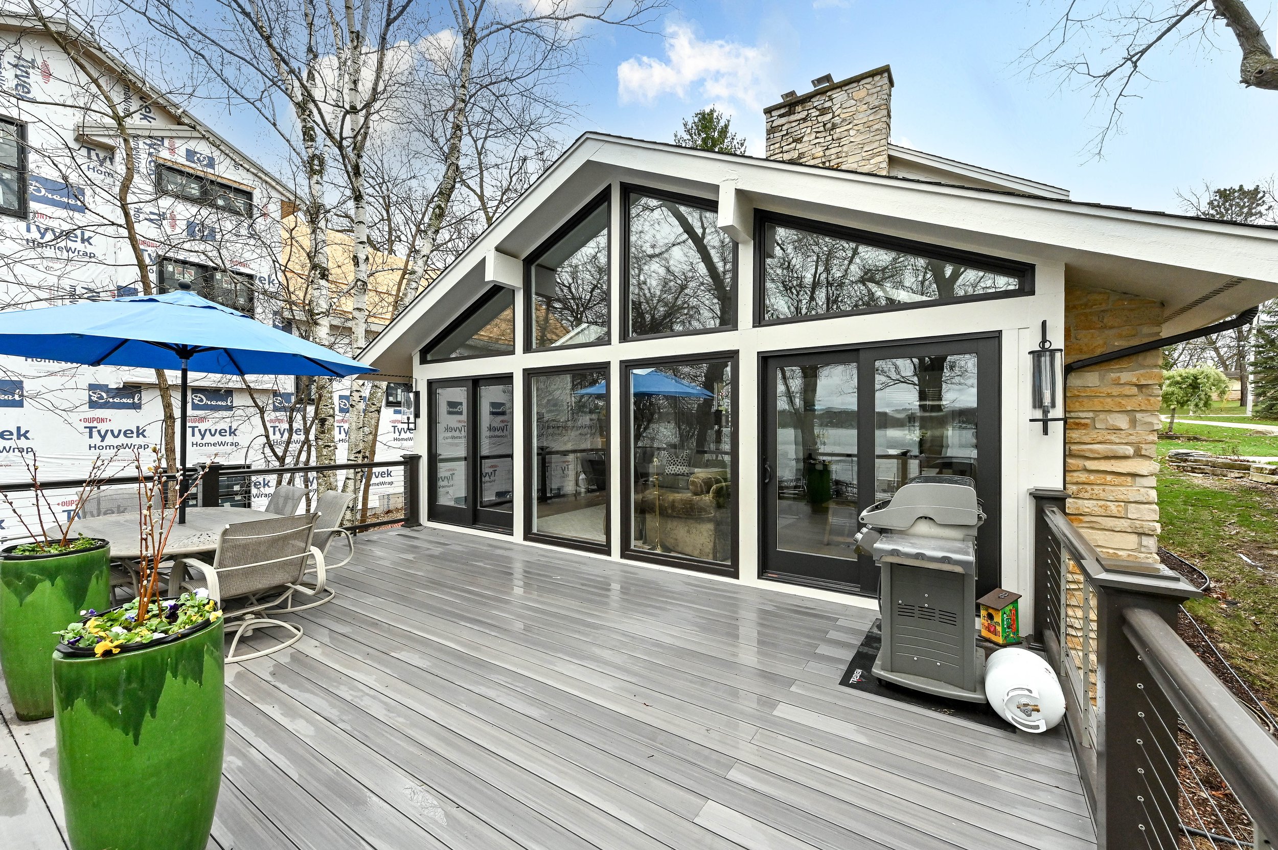 Empty outdoor deck with patio furniture, large green plant pots, a blue umbrella, and a grill, attached to a modern house with large glass windows and a sloped roof.