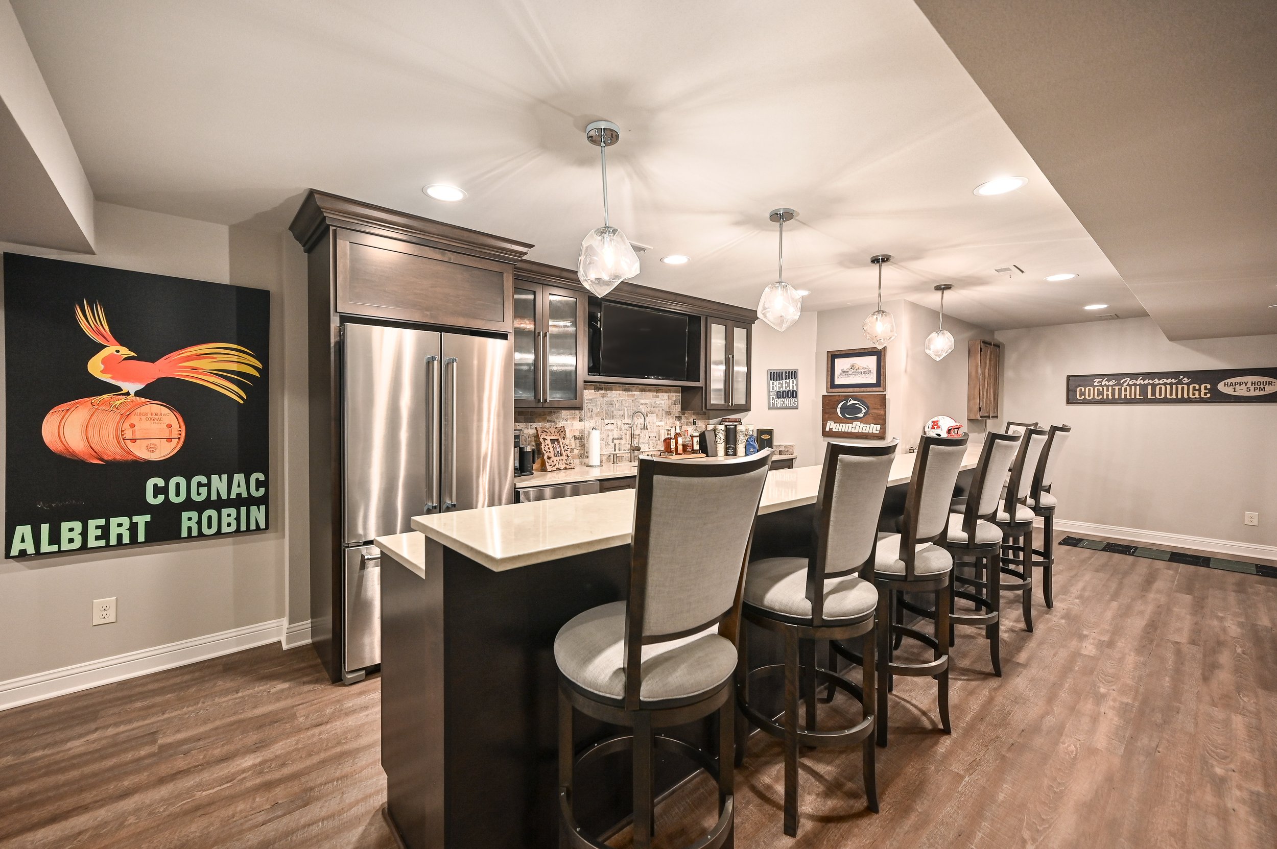 Home bar with a row of upholstered barstools, dark wooden cabinets, a large stainless steel refrigerator, pendant lights, and various signs and decorations on the walls.