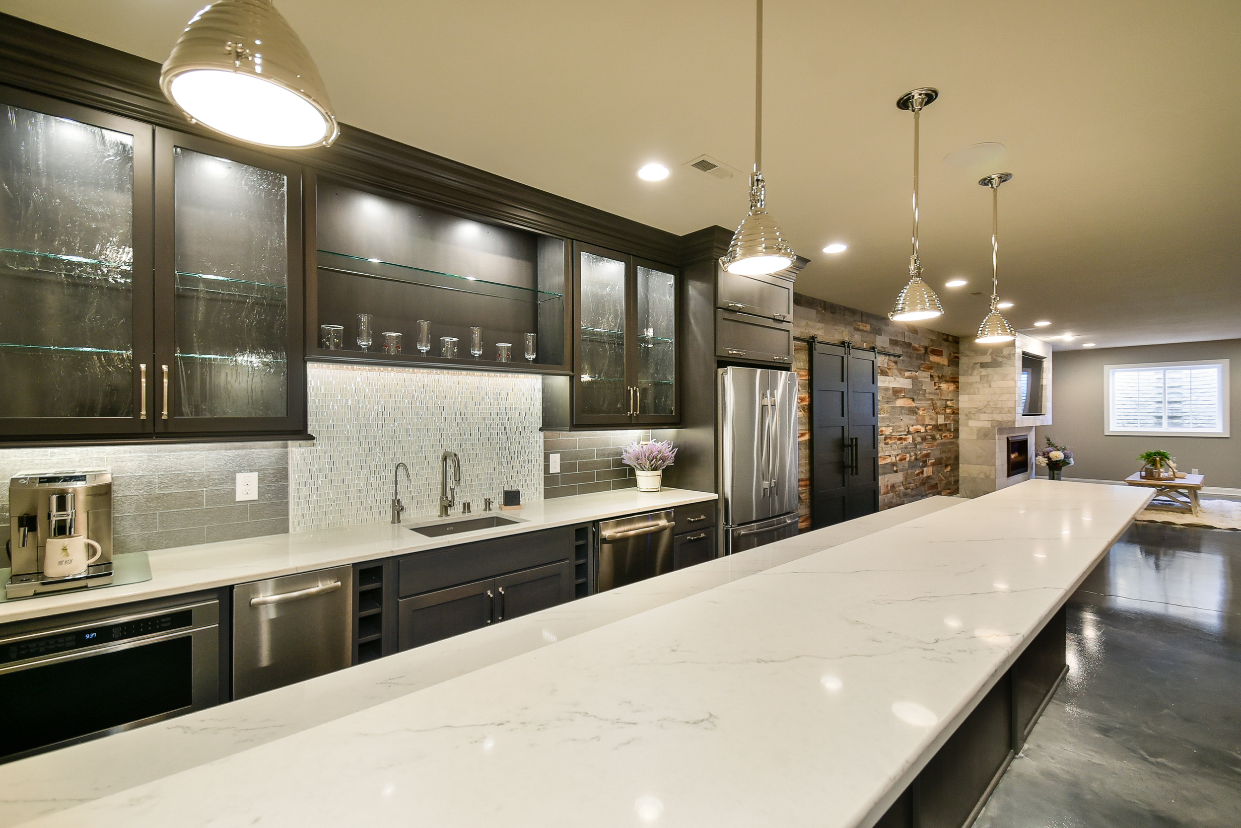 Modern kitchen with dark cabinets, white marble countertop, stainless steel appliances, pendant lights, and a living area with a TV and fireplace.