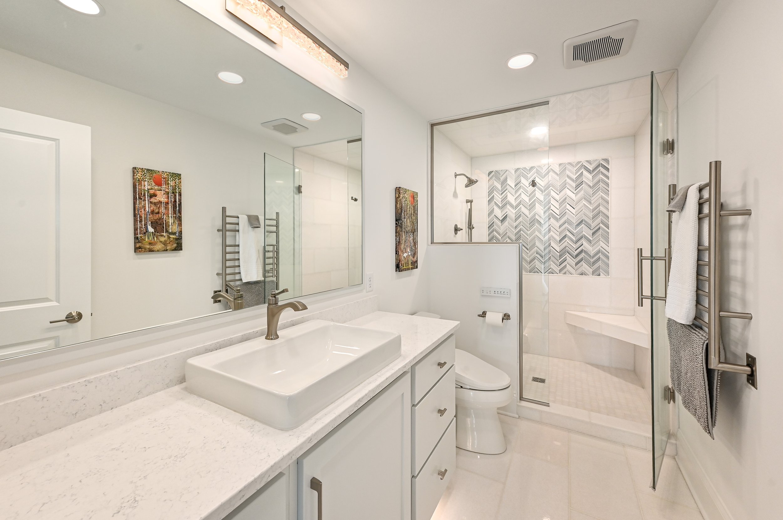 A modern bathroom with a white vanity, rectangular vessel sink, large mirror, and a glass-enclosed shower with patterned tiles.