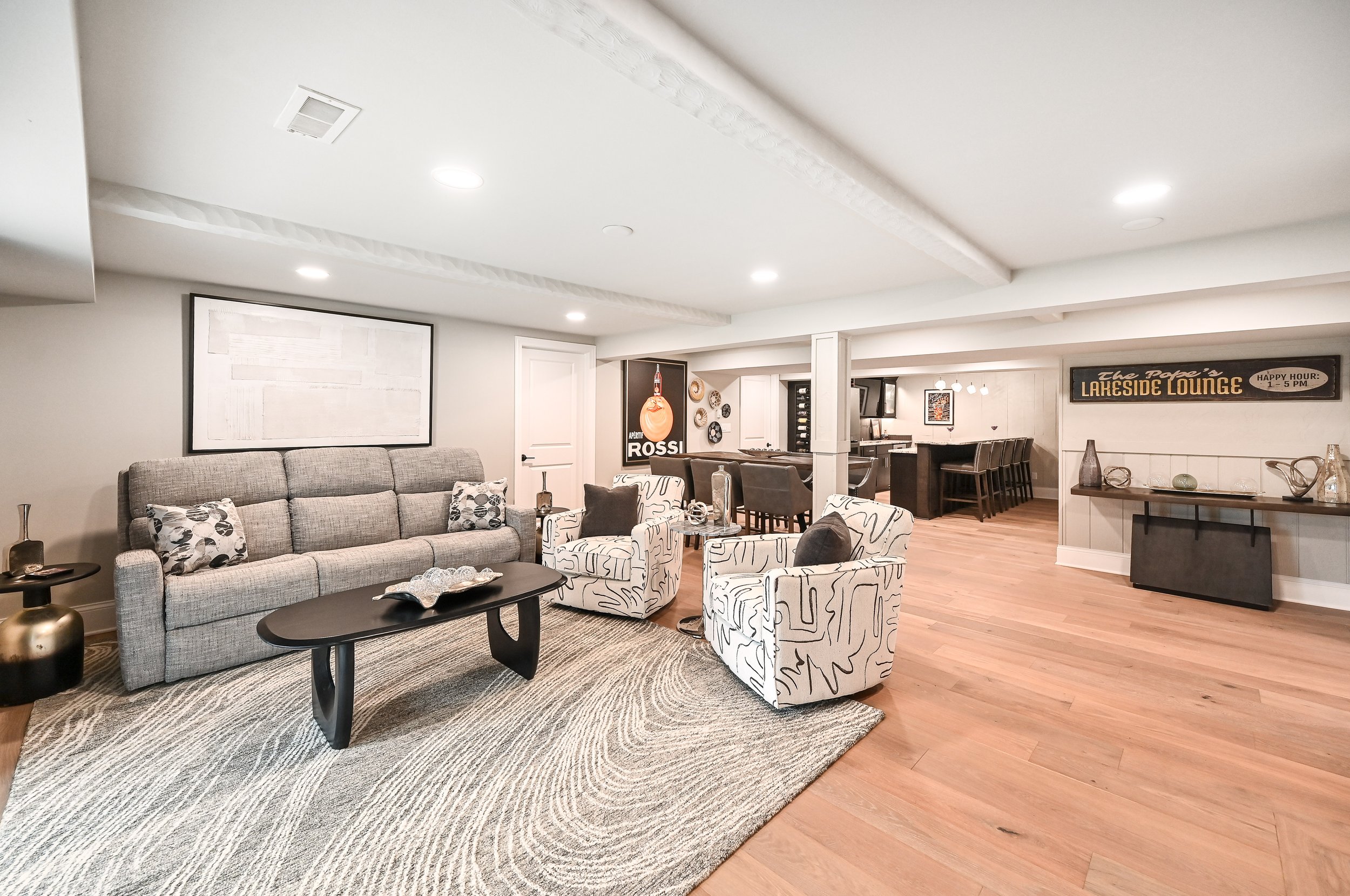 Living room with gray sofa, patterned armchairs, and a rectangular black coffee table on a textured rug. Open kitchen in the background with bar seating and wall decorations including a sign that reads 'Lakeside Lounge'.