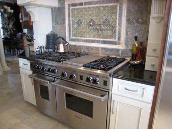 Stainless steel gas stove with oven beneath, on a granite countertop in a kitchen. Above the stove, there is a backsplash with diamond-shaped tiles and a pot filler faucet. To the right, there are bottles of oil and vinegar on the counter. To the lef