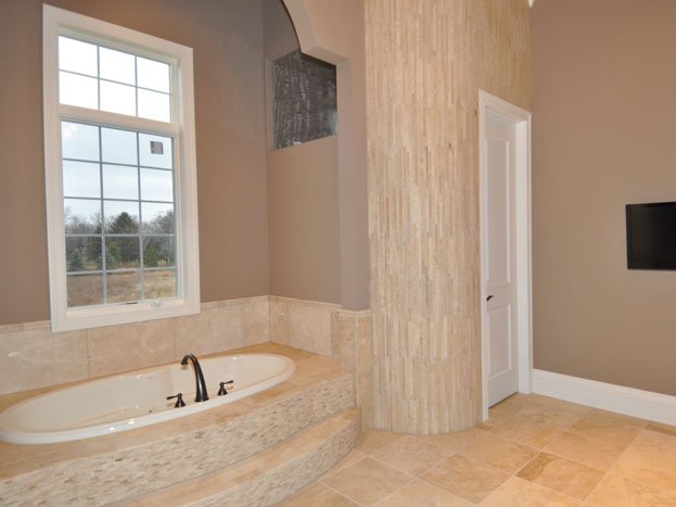 Bathroom with a large bathtub, window, beige tiled walls and floor, a door, and a small wall-mounted TV.