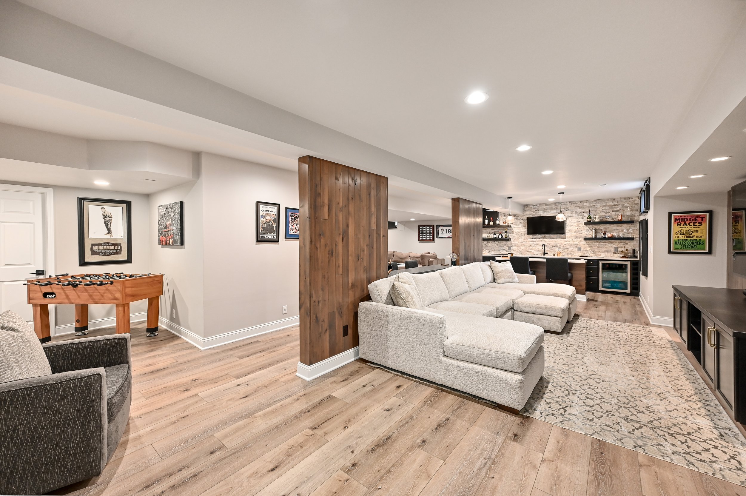 Spacious basement living area with white sectional sofa, flat-screen TV, bar area, framed posters, foosball table, wood accents, and hardwood floors