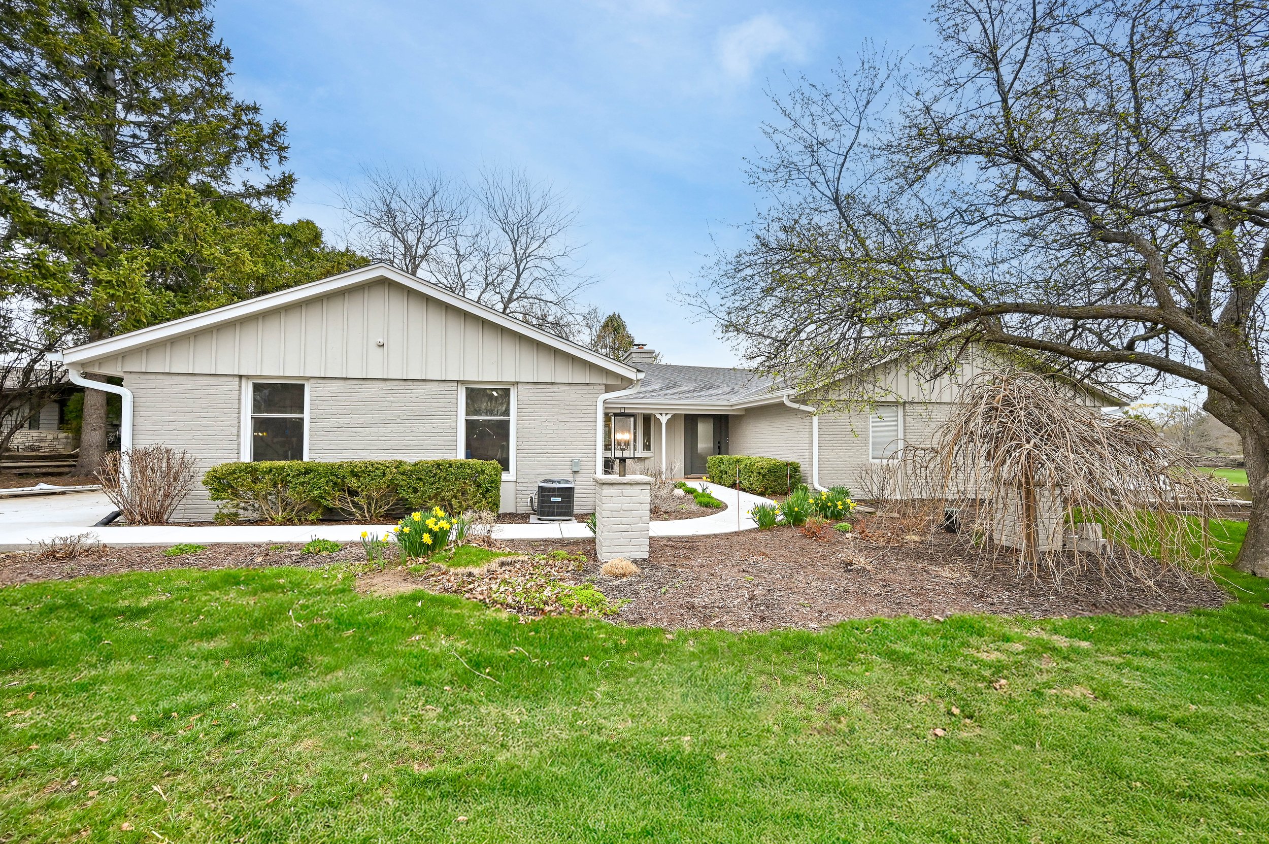 A single-story house with light gray brick exterior, white trim, and a gabled roof. The yard has a green lawn, shrubs, and trees with bare branches. There is a curved walkway leading to the front porch.