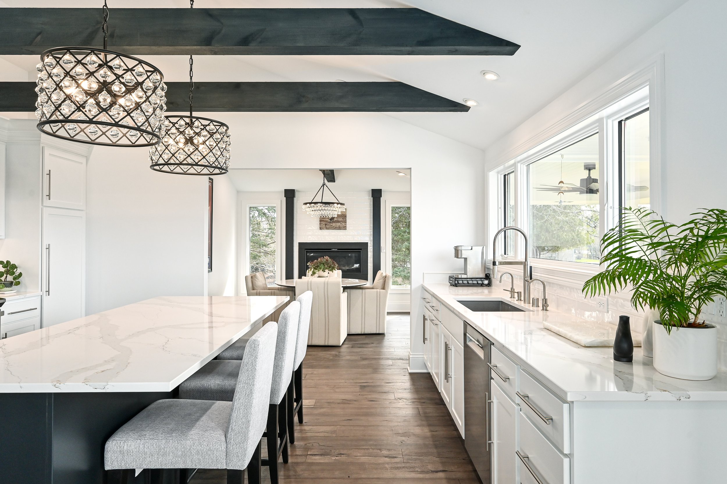 Modern kitchen with white cabinets, marble countertops, large windows, farmhouse sink, black beams on ceiling, and pendant lights, open to living room with fireplace and seating