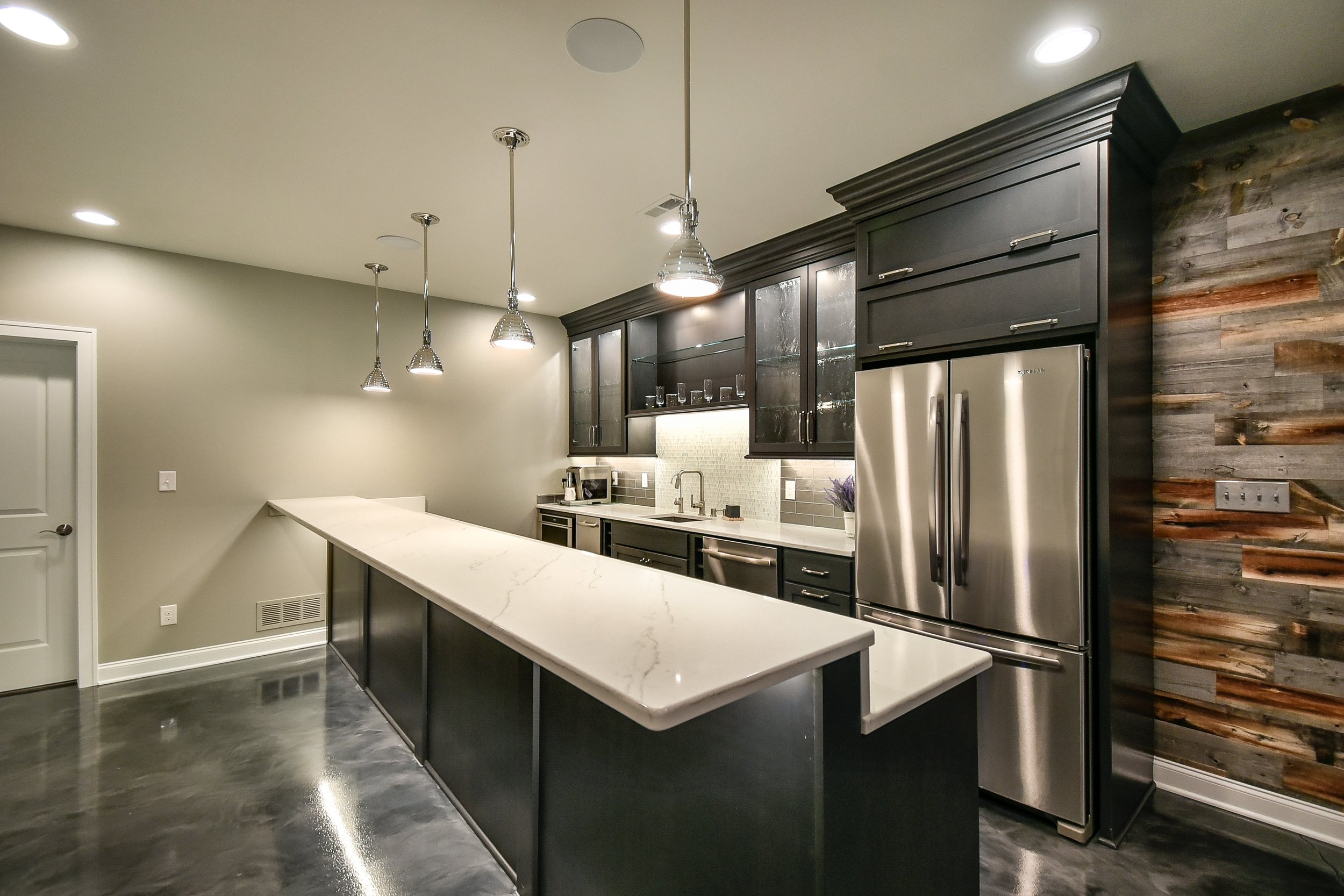 Modern kitchen with dark cabinets, stainless steel refrigerator, white marble island, stone and wood wall accents, pendant lights, and a beige wall.