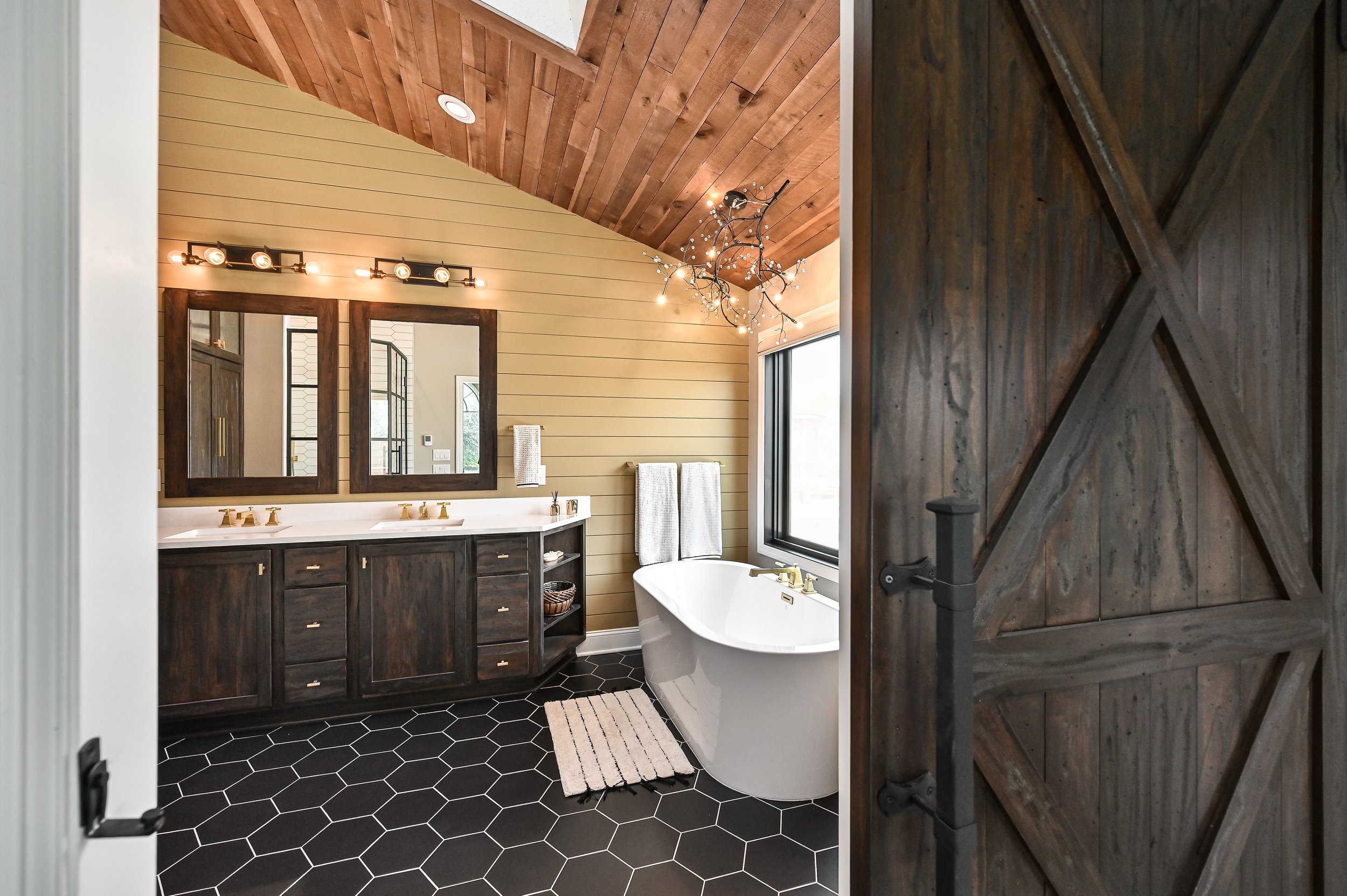 Modern rustic bathroom with a wooden sliding barn door, black hexagonal floor tiles, a large window, a freestanding bathtub, a double vanity with mirrors, and decorative lighting and chandelier.
