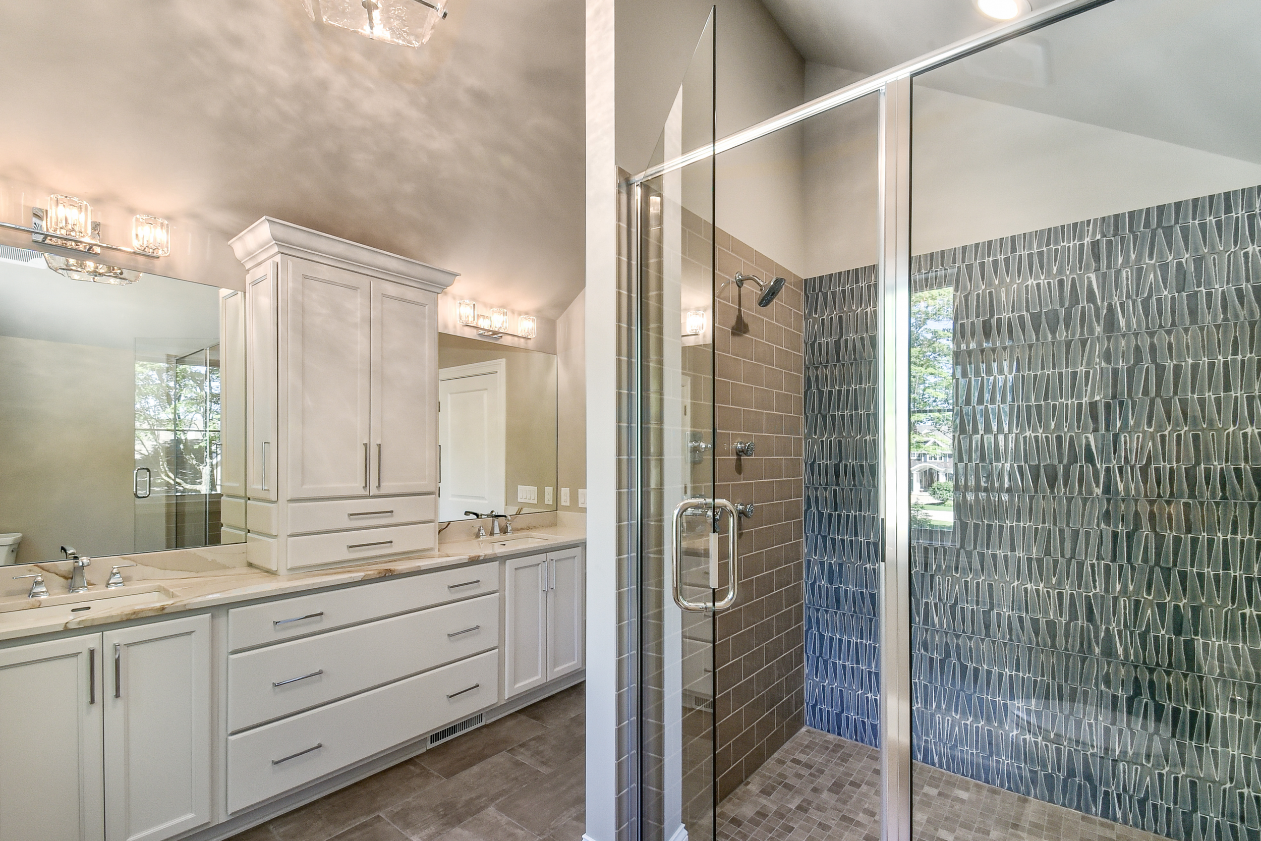 Modern bathroom with double vanity sinks, white cabinetry, and large mirror; walk-in shower with glass enclosure, brown brick, and blue patterned wall tiles; bathroom lighting fixtures; window with outdoor greenery visible.