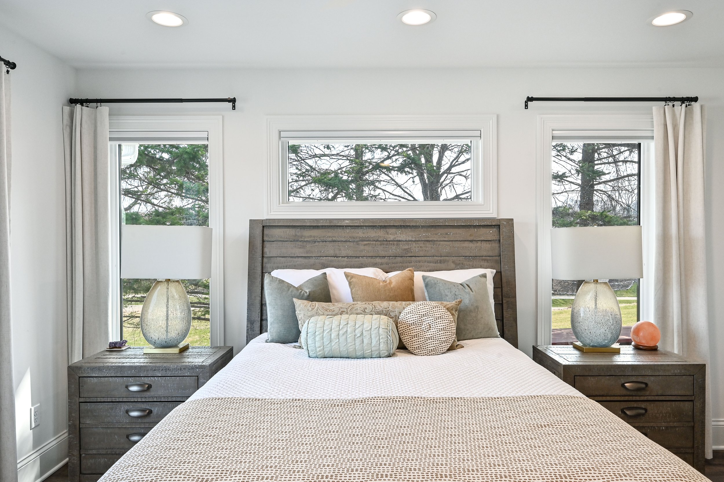 A bedroom with a wooden headboard, white bedding, and multiple decorative pillows. Two nightstands with lamps and decorative items are on each side of the bed. Three windows with curtains and a view of trees outside.