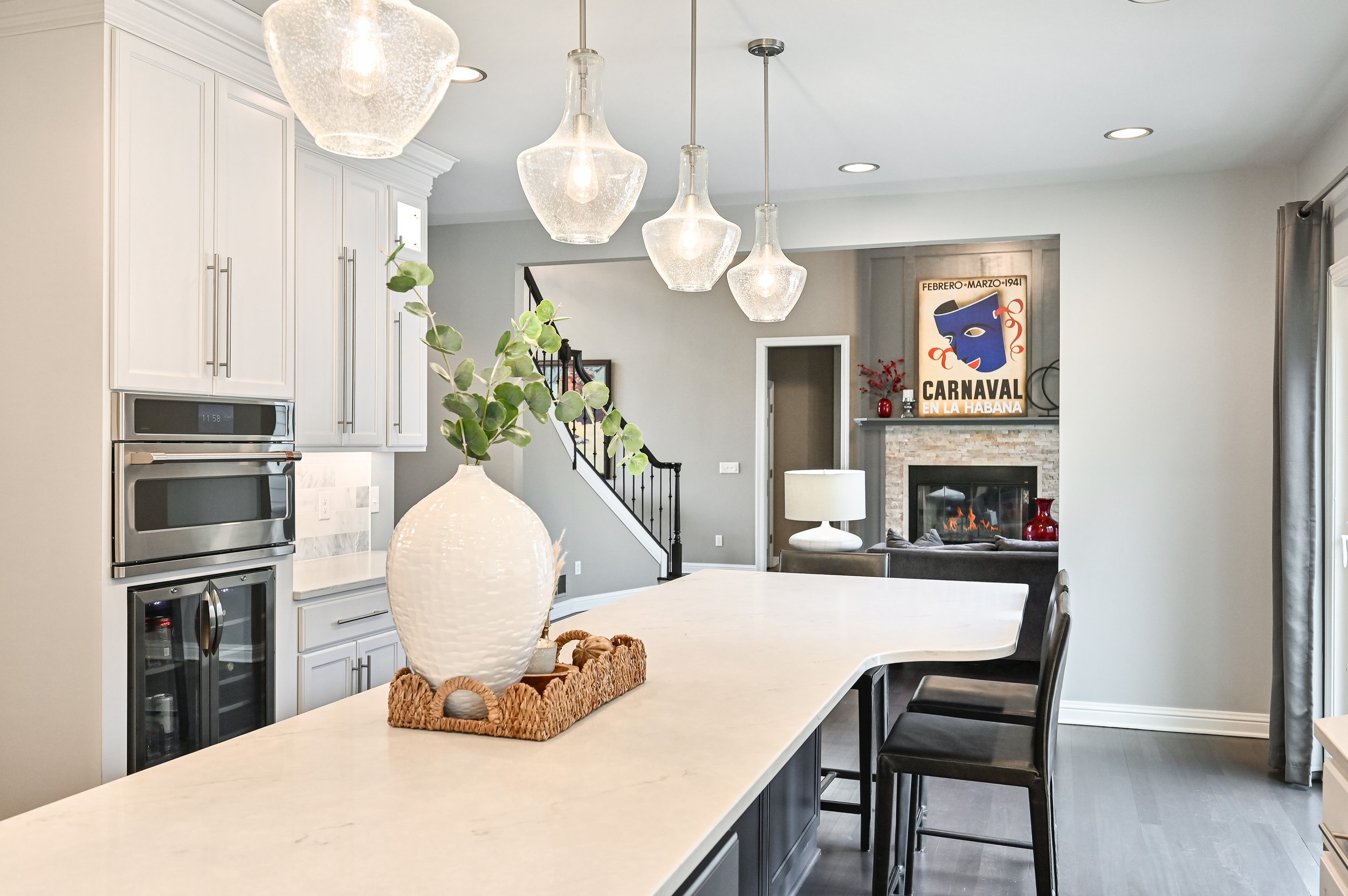 Modern open-concept kitchen with white cabinets, a marble countertop island, large glass pendant lights, a decorative vase with greenery, and a living area with a fireplace, artwork, and seating.