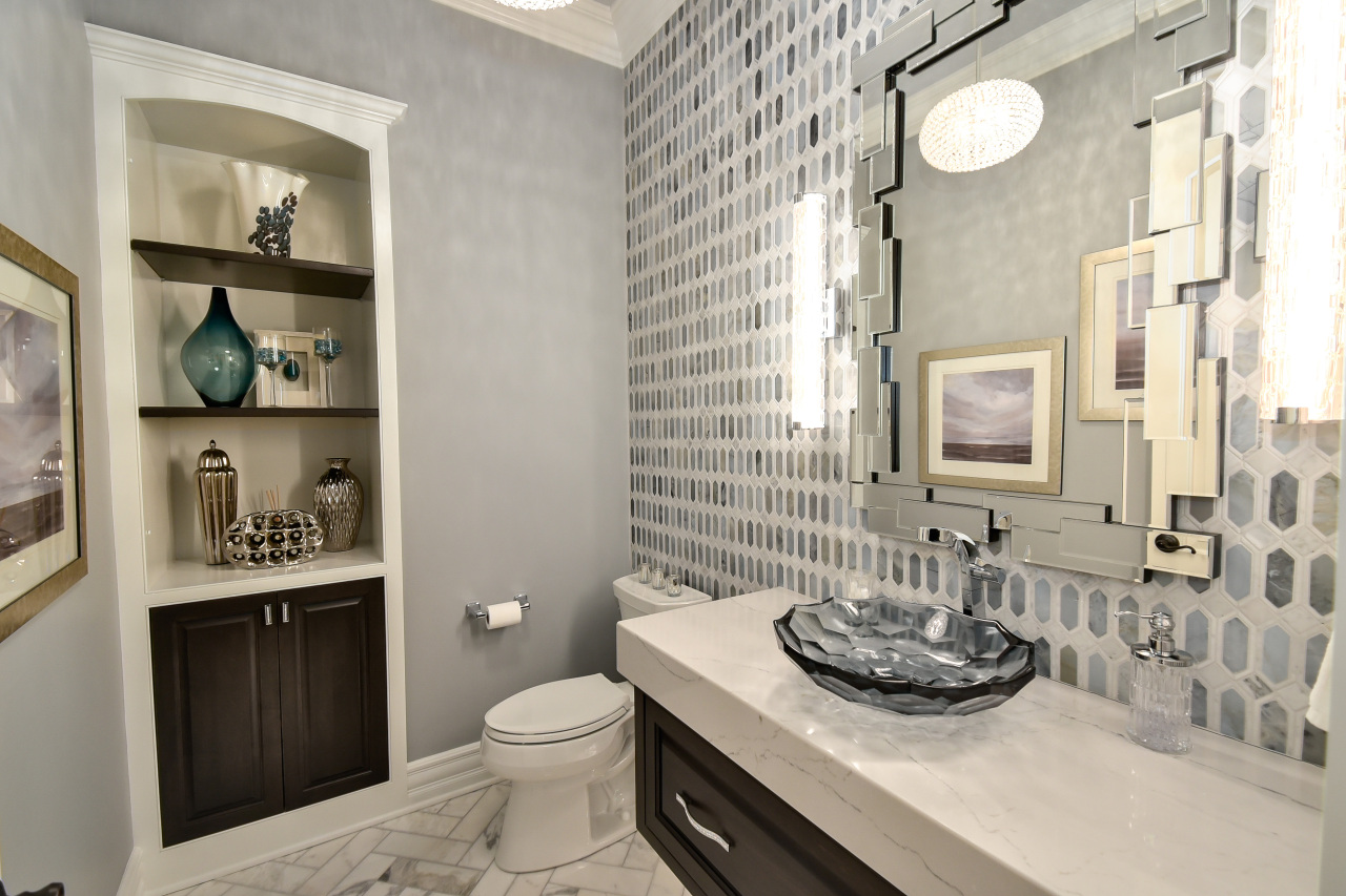 Modern bathroom with patterned tile wall, large mirror, pedestal sink with vessel basin, decorative items on built-in shelves, toilet, and framed artwork.