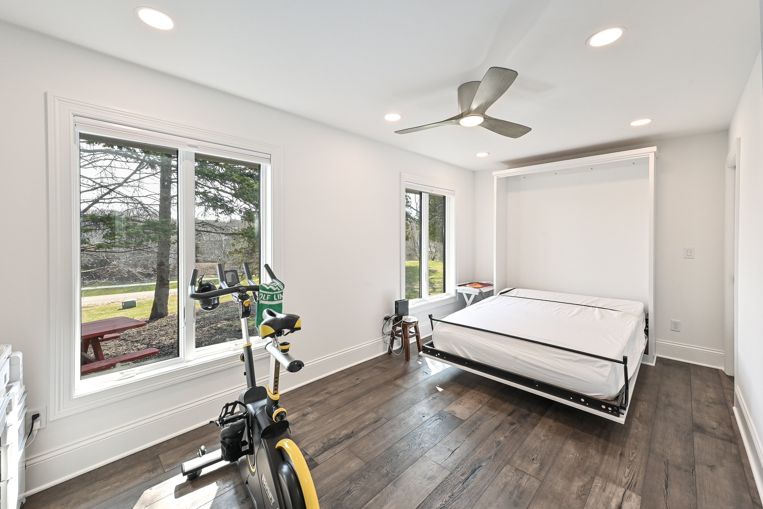 A minimalistic bedroom with white walls and dark hardwood floors, containing a bed without bedding, a stationary exercise bike, large windows with views of trees and a lawn, a ceiling fan, and a small side table.