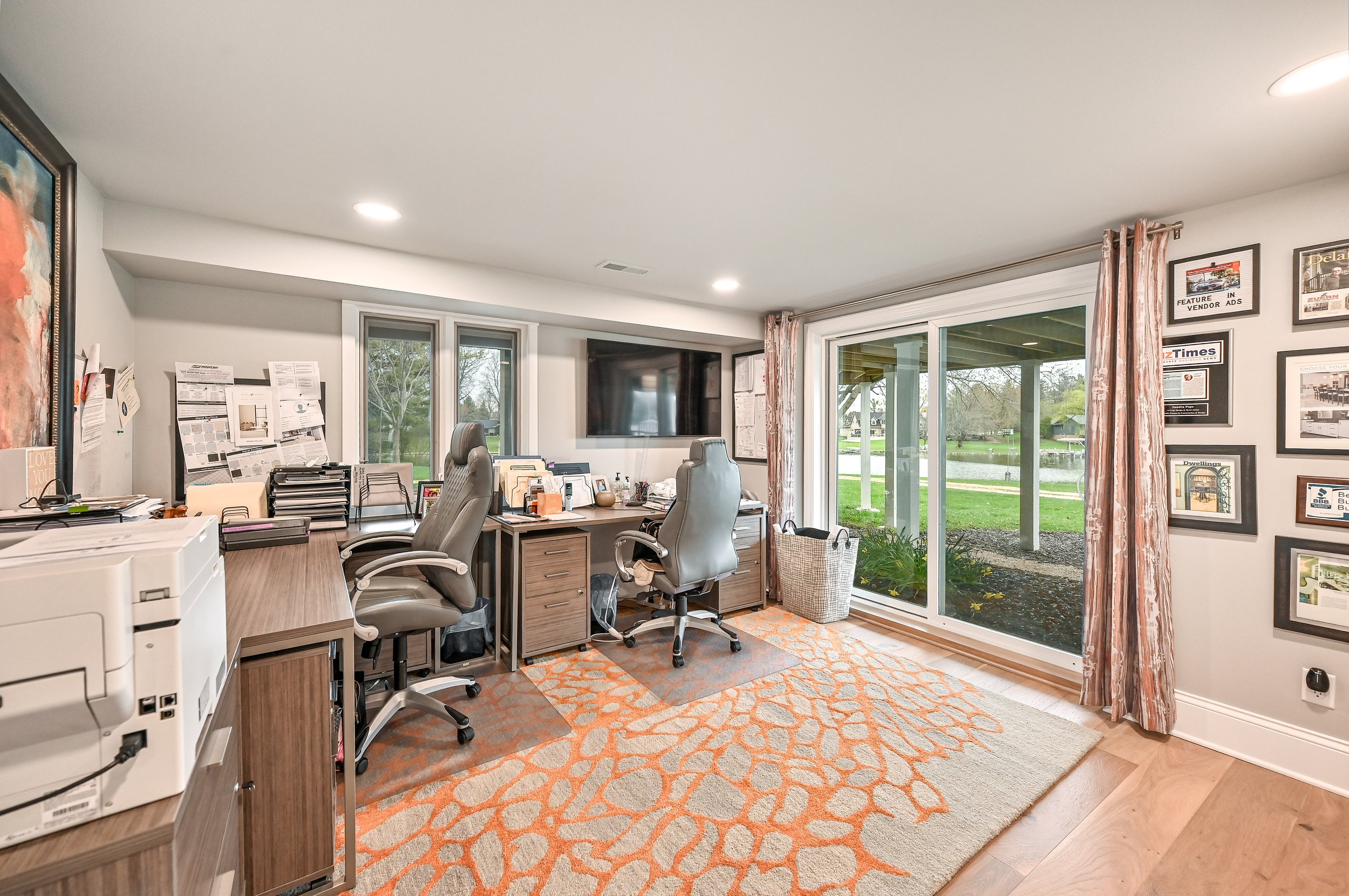 Home office with two desks, two chairs, a wall-mounted TV, and a sliding glass door with curtains, overlooking a backyard with trees and a lake.