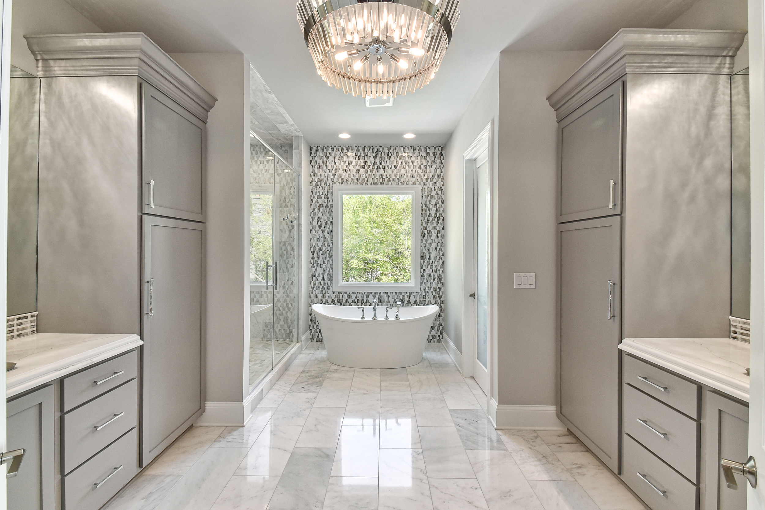 Modern bathroom with gray cabinets, marble tile floor, a freestanding bathtub underneath a window, a walk-in shower with glass door, and decorative tiled accent wall behind the bathtub.