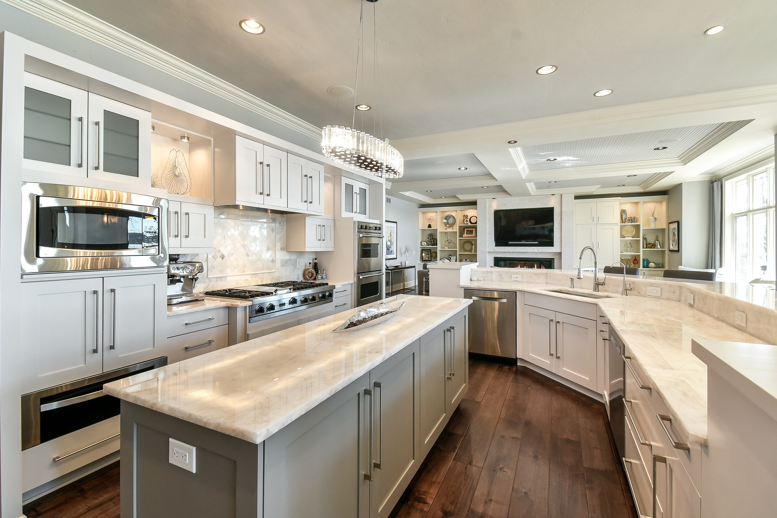 Modern kitchen with white cabinets, marble countertops, stainless steel appliances, and hardwood flooring. There's a central island with a support base, a built-in microwave, and a large window letting in natural light.